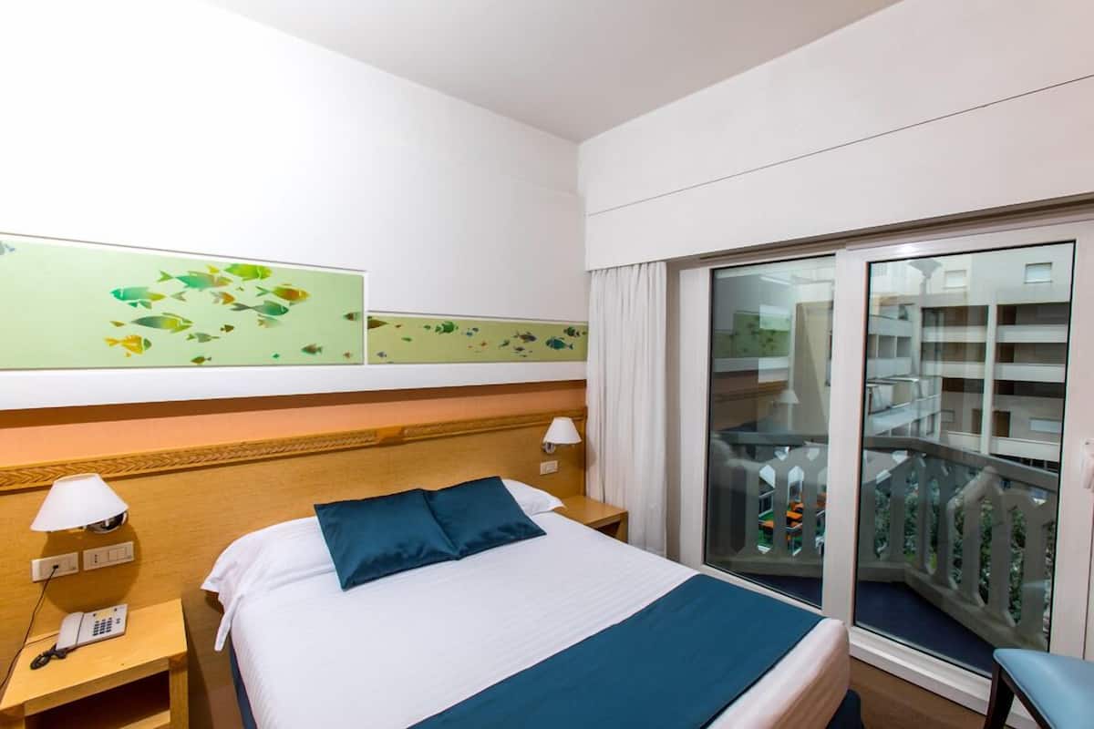 A hotel room with a bed, a nightstand with a phone, a window with a view of a balcony, and artwork on the wall.