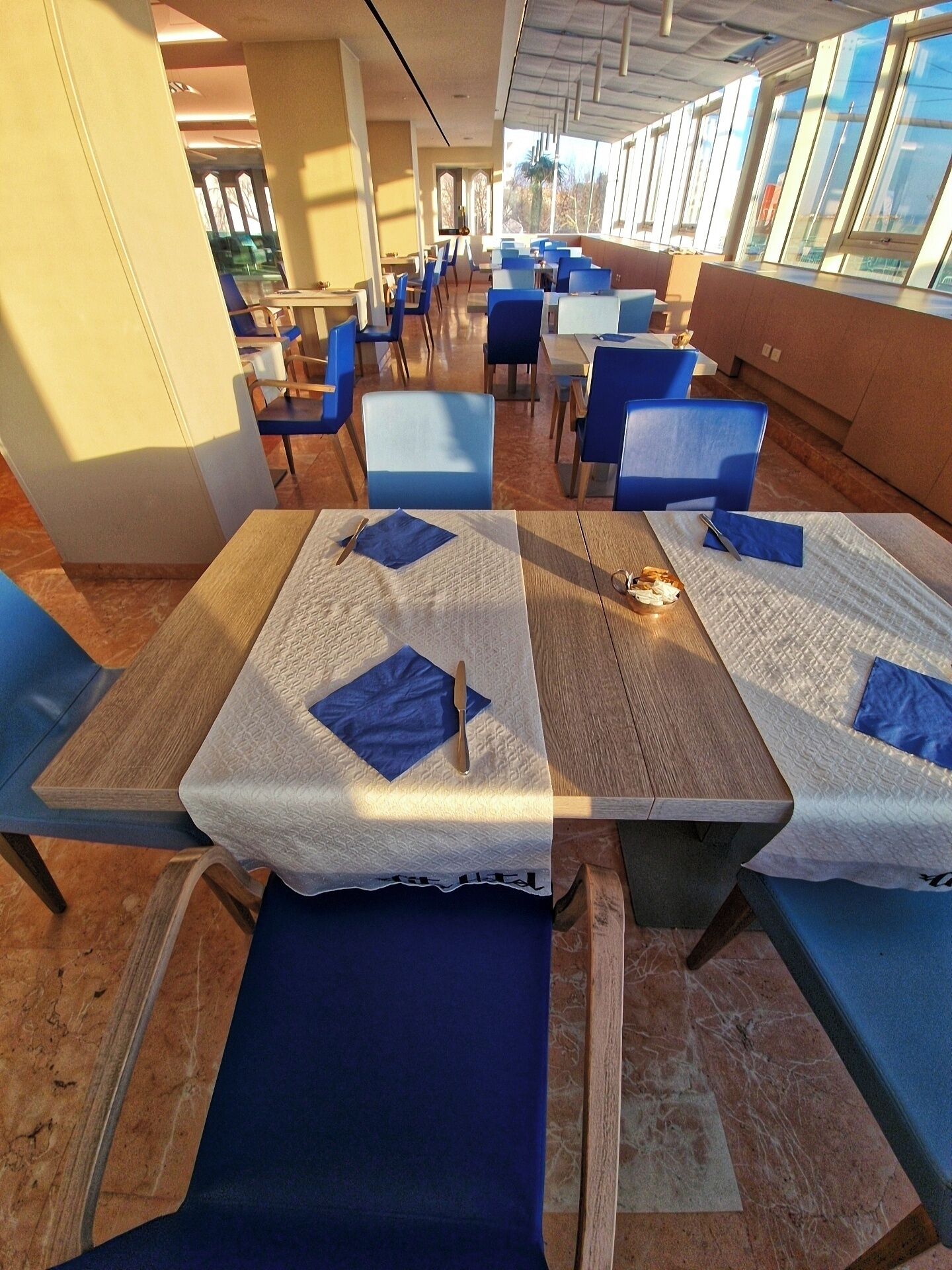 A restaurant with tables covered with white tablecloths and blue napkins, blue chairs, and a view of the outdoors through large windows.