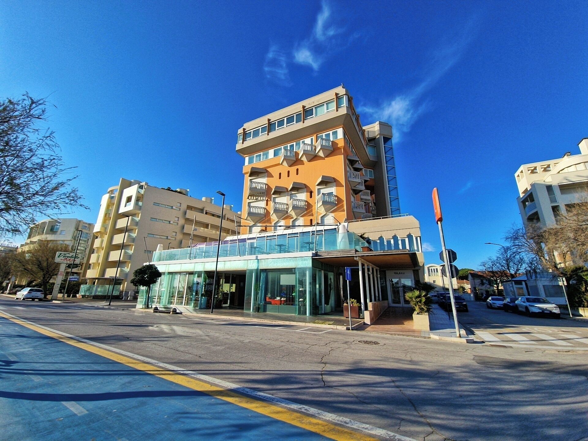 A modern hotel with a glass facade, multiple balconies, and a pedestrian walkway.