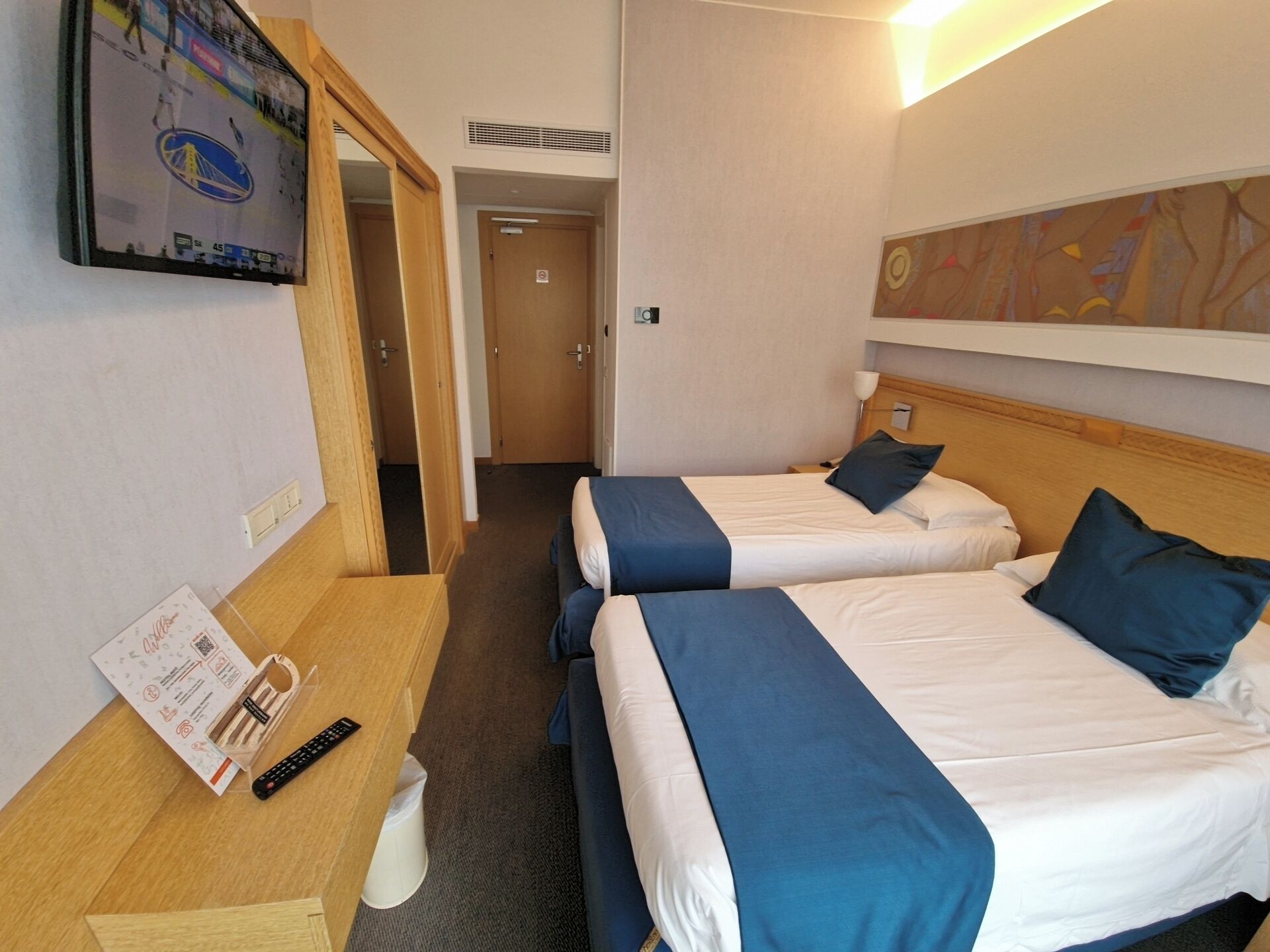 Ein Hotelzimmer mit zwei Betten, einem an der Wand befestigten Fernseher, einer Fernbedienung und einer Tür zu einem weiteren Raum.