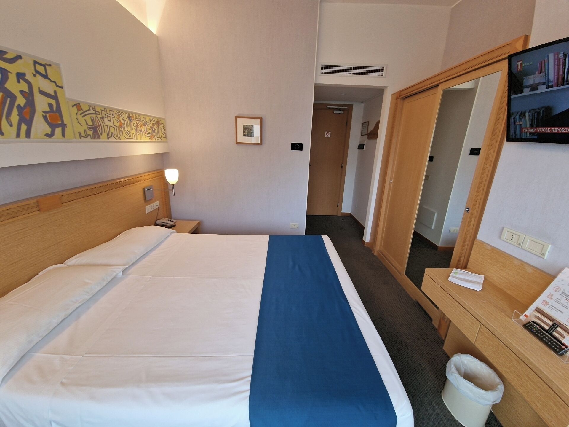 Quarto de hotel com cama, escrivaninha com cesto de lixo, espelho e parede com obras de arte.