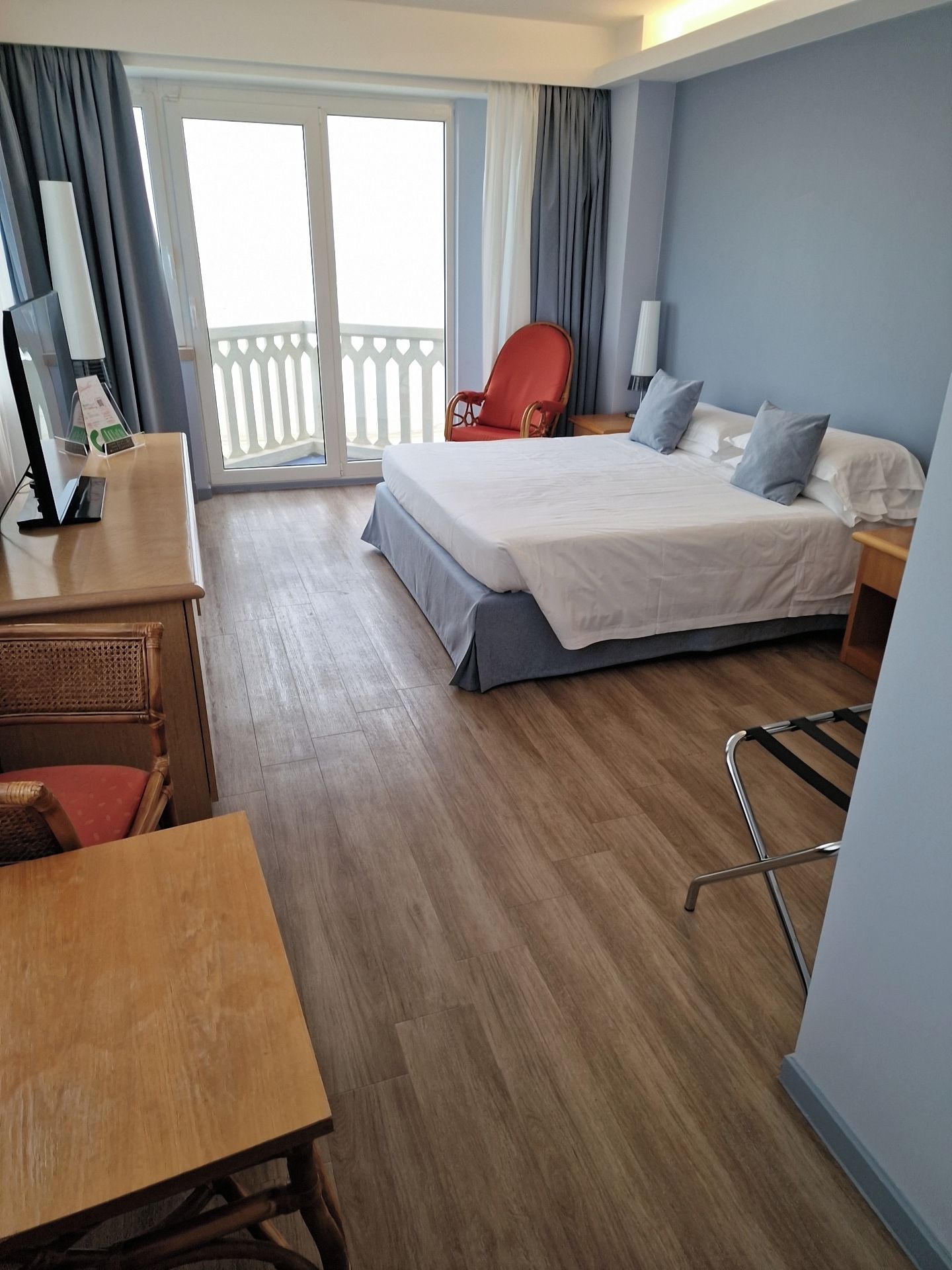 Ein Hotelzimmer mit Bett, Schreibtisch, Stuhl, Fernseher und einem Balkon mit Vorhängen.