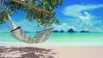 A hammock tied to a tree branch over a beach with clear turquoise water and distant islands.