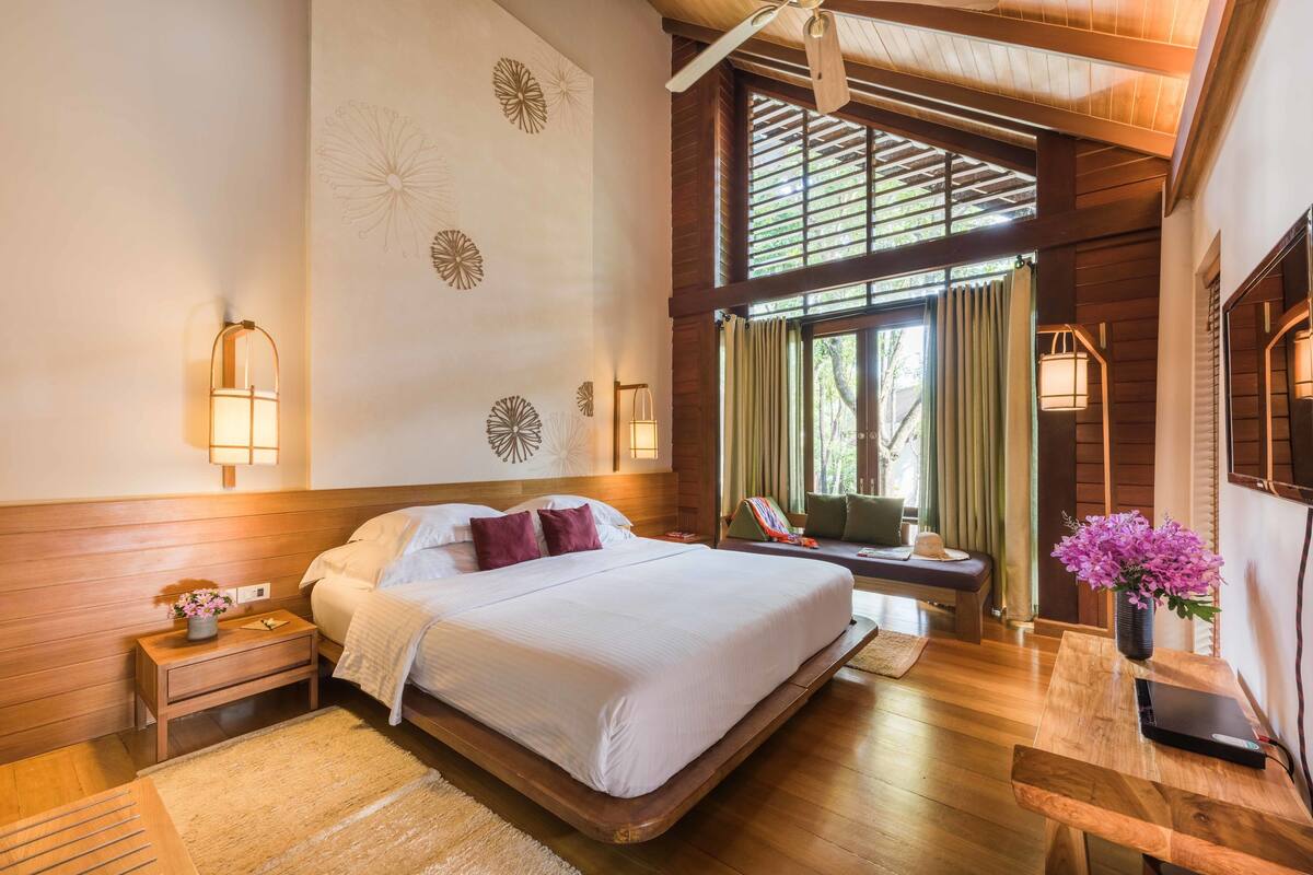 A spacious bedroom with a large bed, wooden headboard, and a view of the outdoors through a window with curtains.