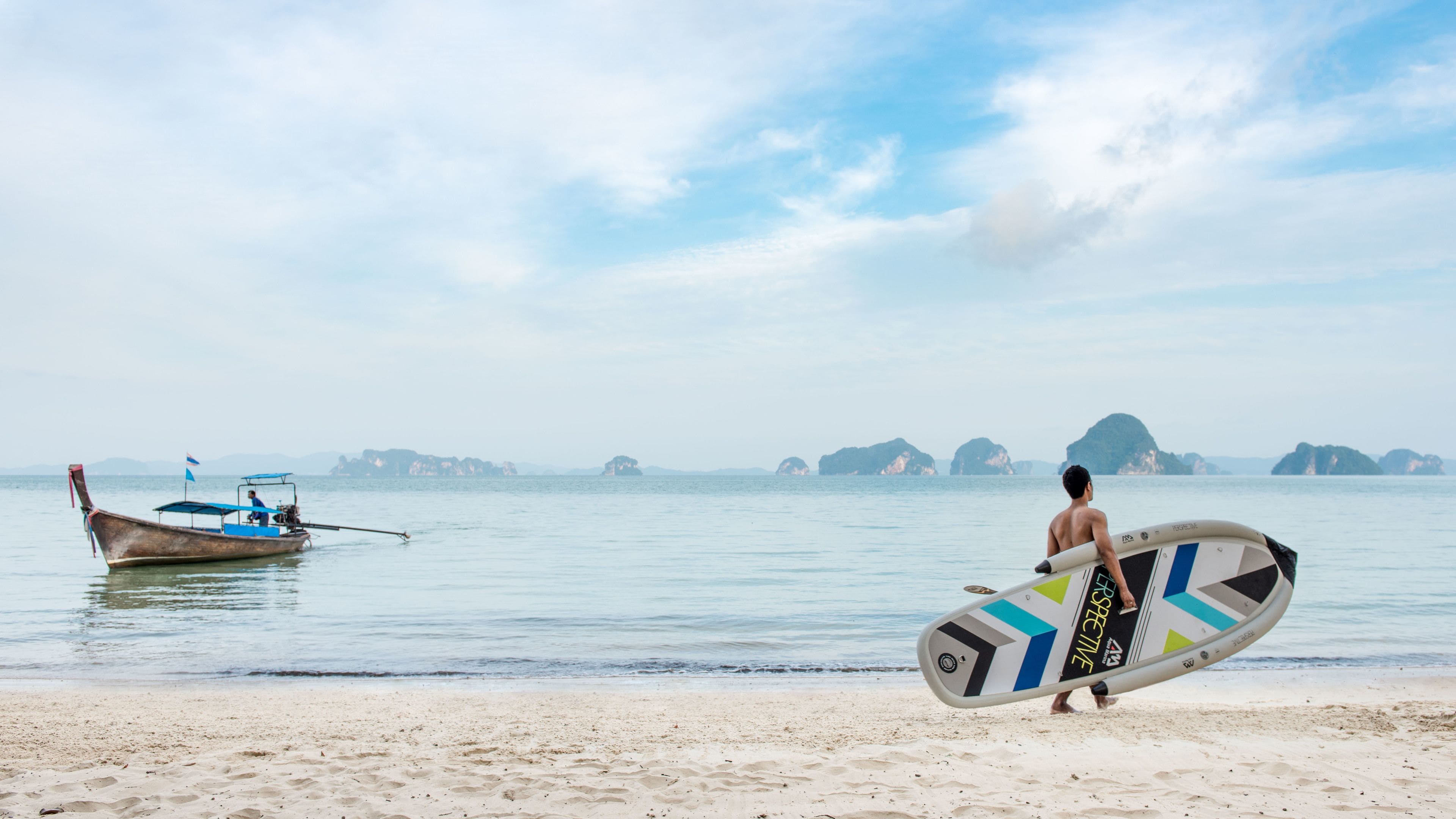Une plage où l’on voit une personne portant une planche de surf, un bateau sur l’eau et des îles au loin.