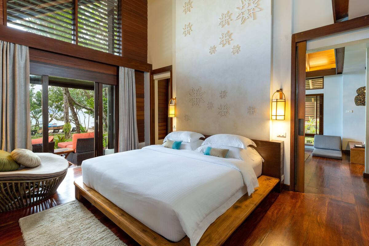 A well-lit bedroom with a large bed, a wicker chair, and a view of the outdoors.