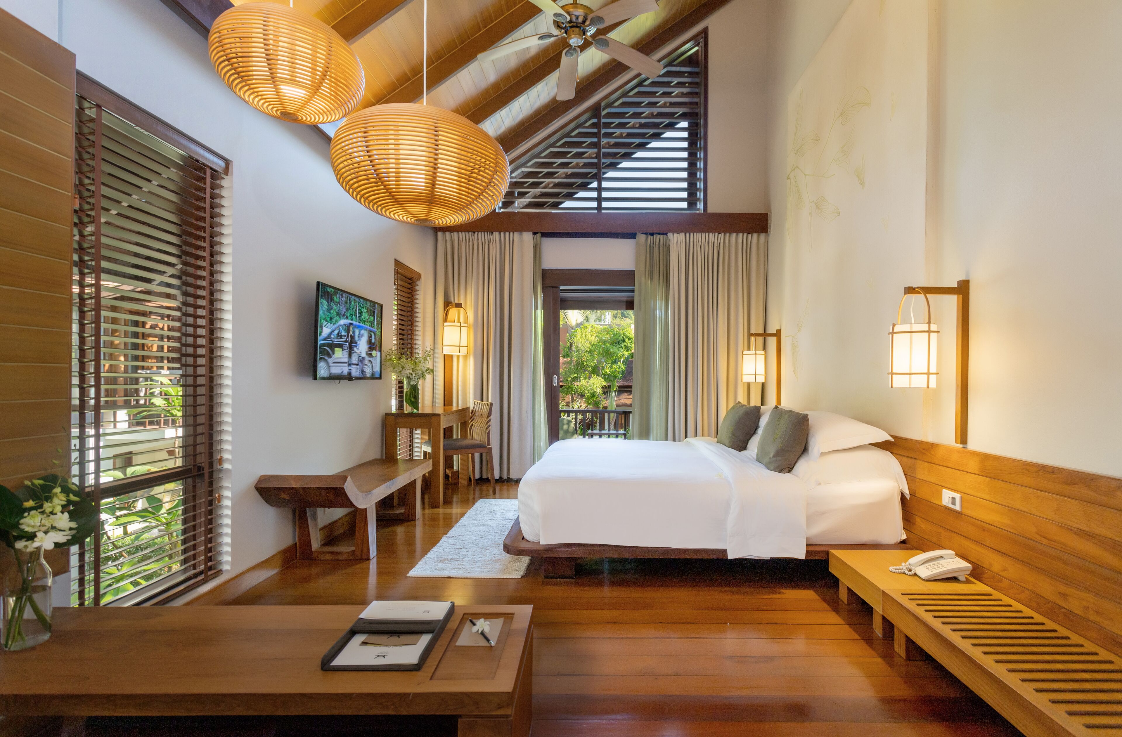 A hotel room with a large bed, wooden furniture, a desk, and a view of greenery outside.
