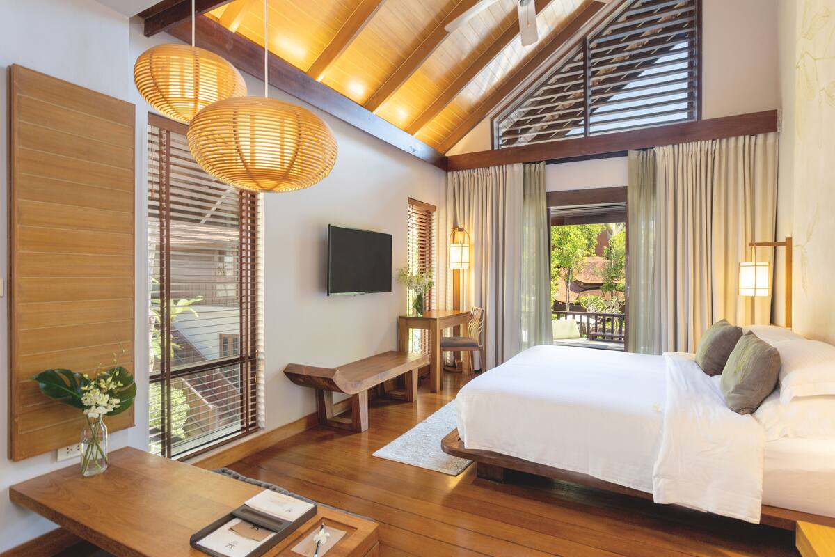 A bedroom with a large bed, wooden flooring, a bench, a TV, and a view of the outdoors.