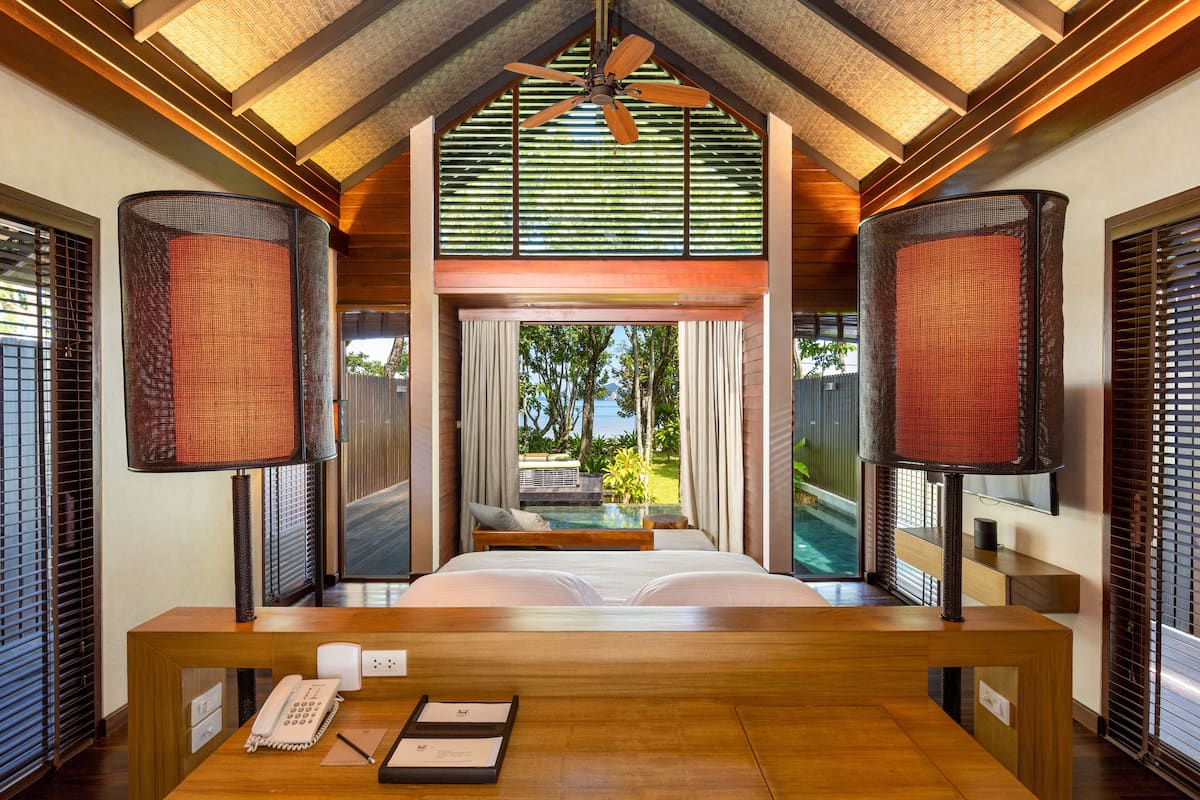 A bedroom with a wooden bed, a desk, a telephone, and a window with a view of a pool and greenery.