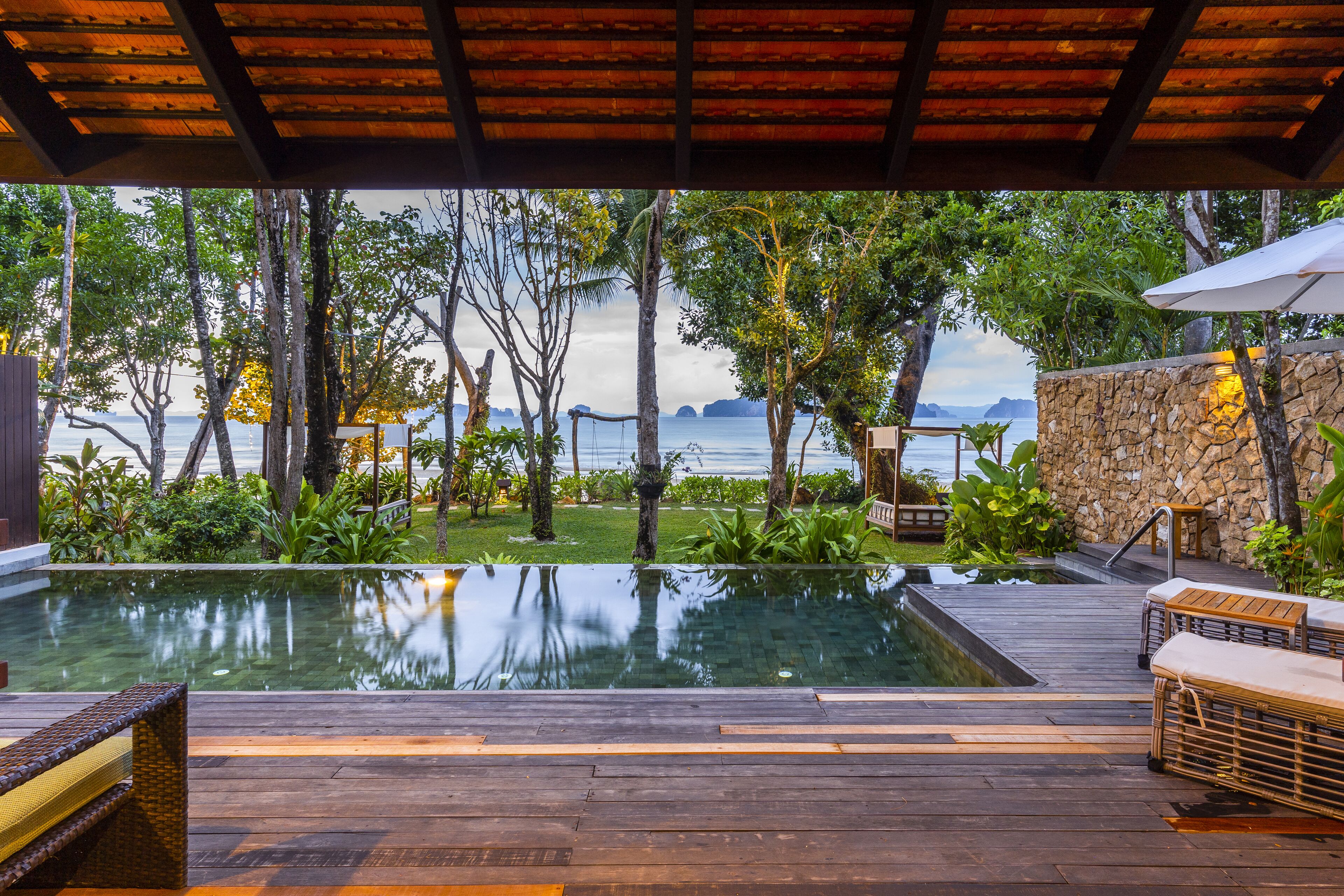An infinity pool with a wooden deck and lounge chairs, surrounded by lush greenery and a stone wall, overlooking a body of water.