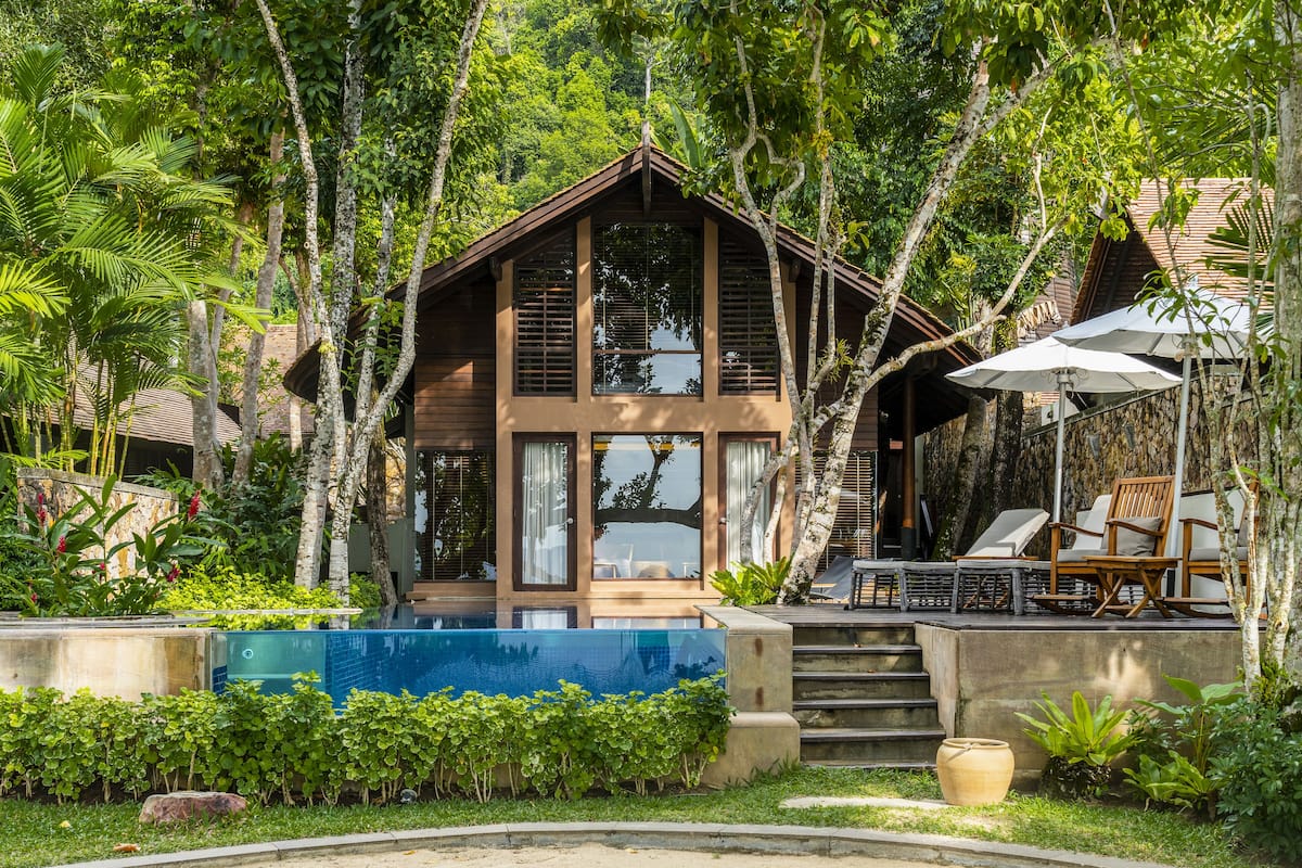 A wooden villa with a swimming pool, surrounded by lush greenery and a deck with outdoor furniture.