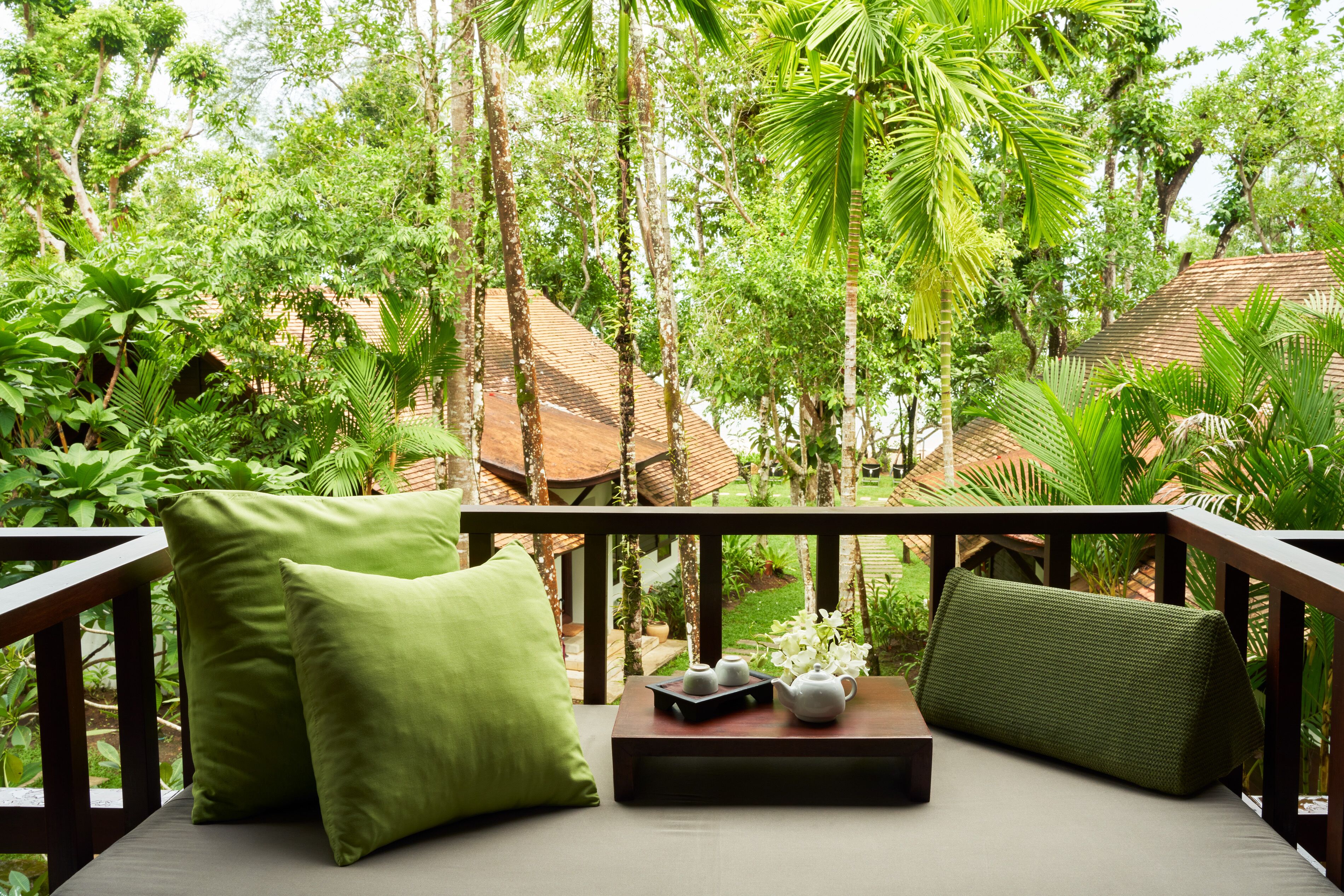 A balcony with a cushioned seating area, a small table with teapots, and lush greenery in the background.