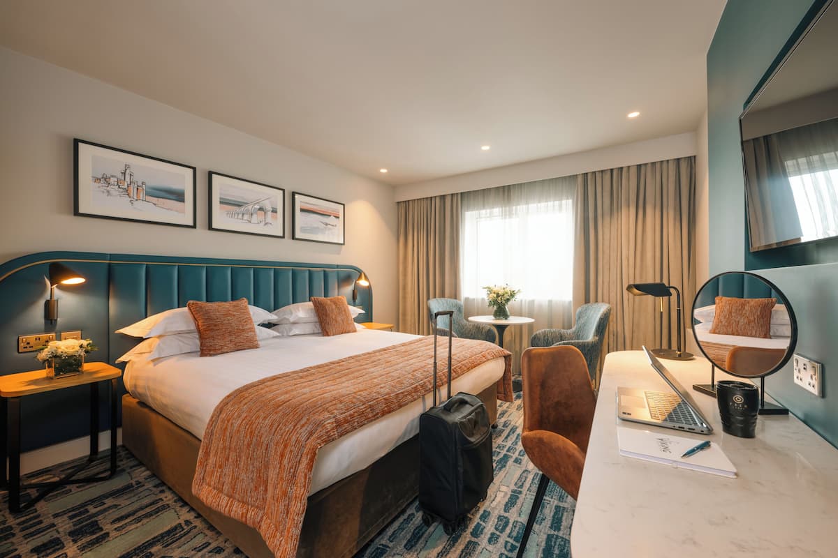 Une chambre d’hôtel avec un grand lit, un bureau avec un ordinateur portable, une chaise, une petite table avec un vase et un miroir.