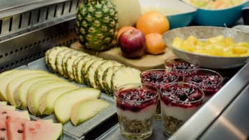 A breakfast buffet with sliced fruits, a melon, a bowl of pineapple chunks, and glasses of fruit salad.