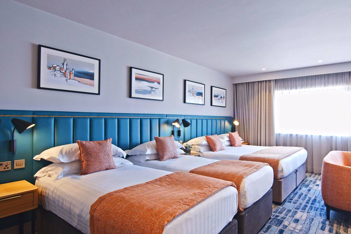 Une chambre d’hôtel avec deux lits, une tête de lit bleue, une grande fenêtre avec des rideaux et une chaise.