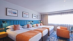 Une chambre d’hôtel avec deux lits, une tête de lit bleue, une grande fenêtre avec des rideaux et une chaise.