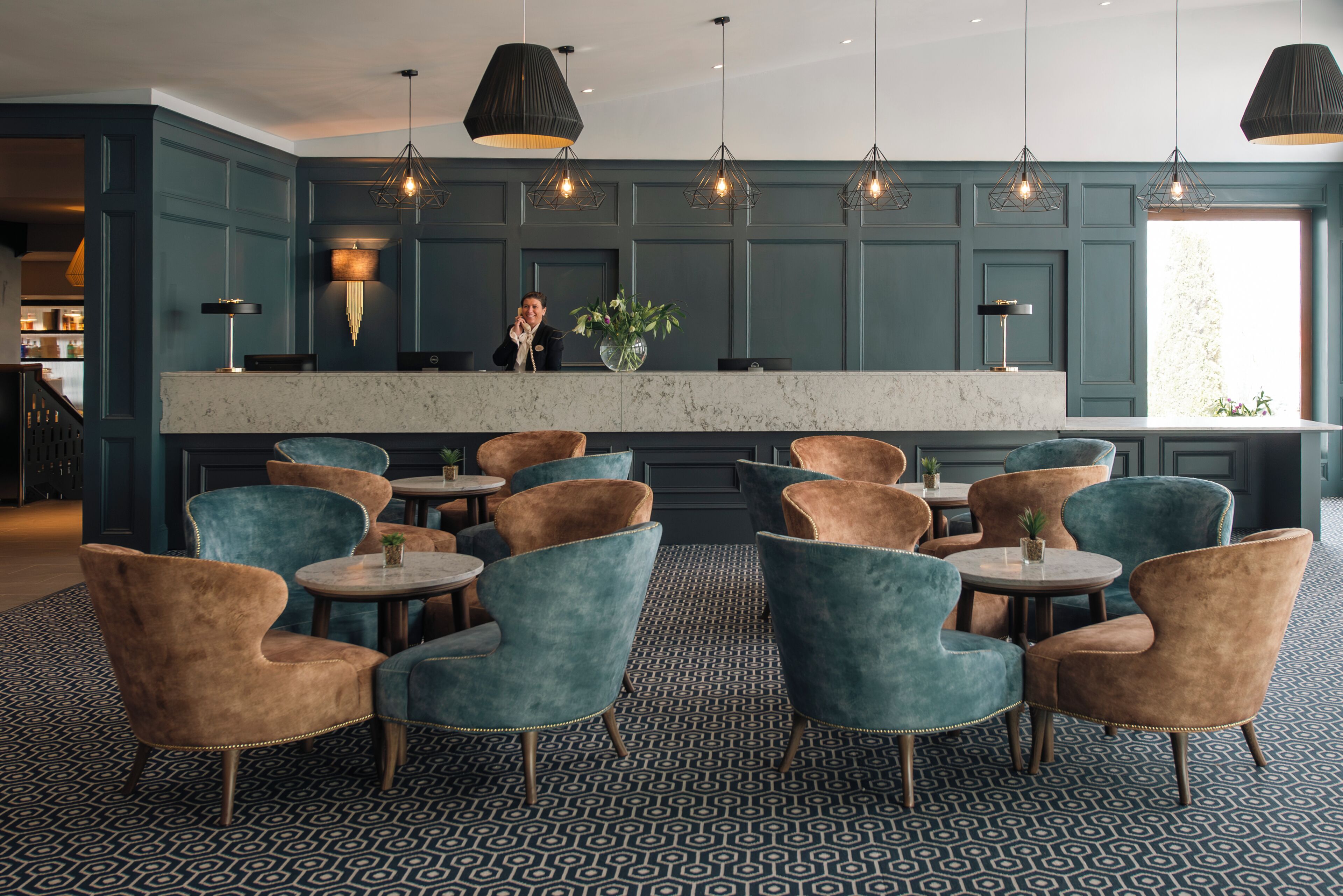 Un vestíbulo de hotel con paredes color turquesa, un mostrador de recepción de mármol y varias zonas de estar con mesas y sillas redondas.