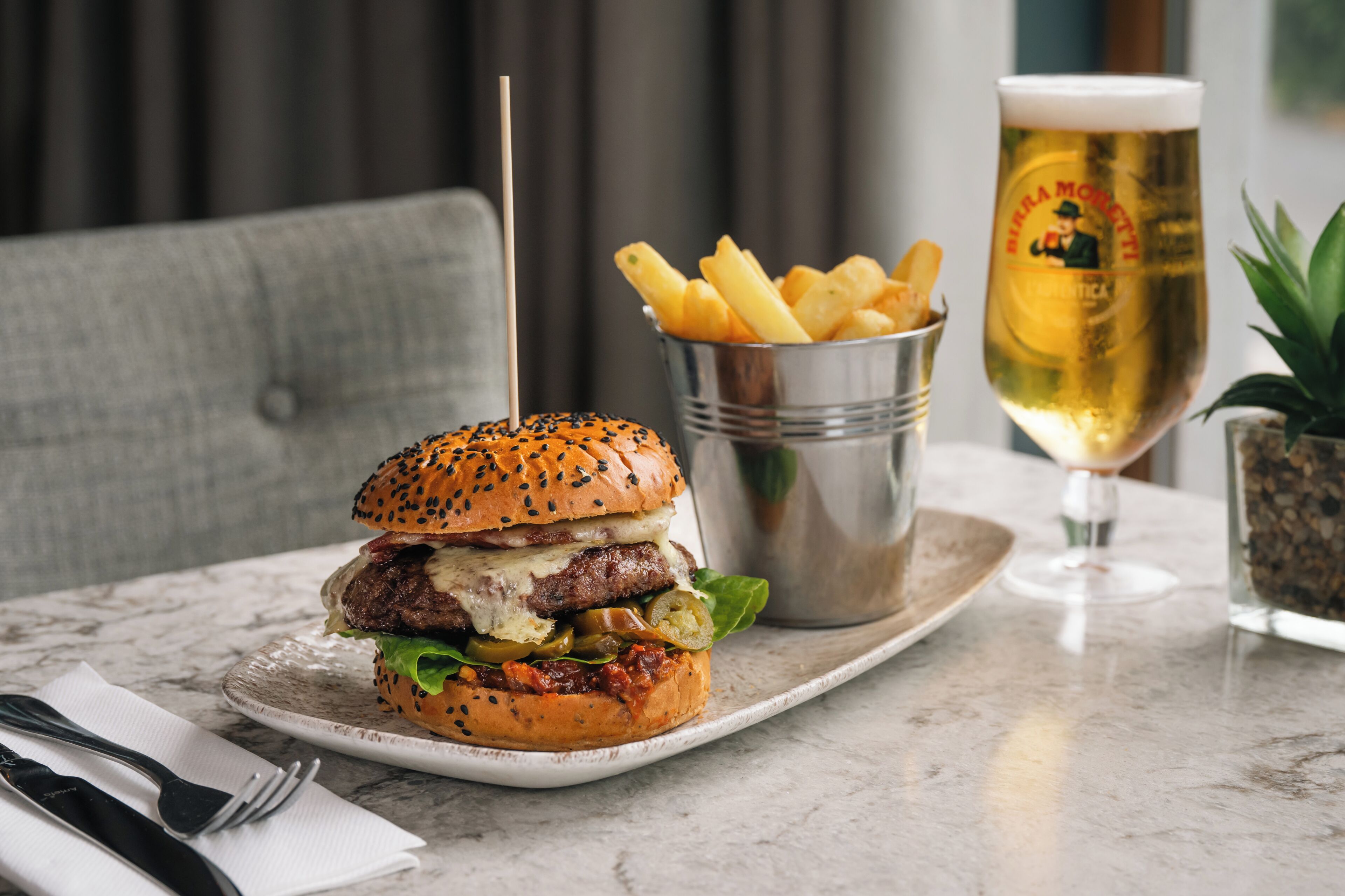 A burger with a sesame seed bun, a glass of beer, and a small metal container of fries on a marble table.