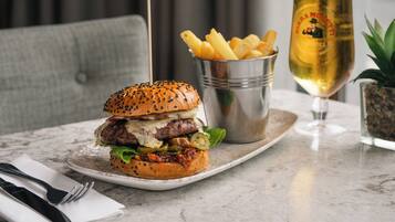 A burger with a sesame seed bun, a glass of beer, and a small metal container of fries on a marble table.