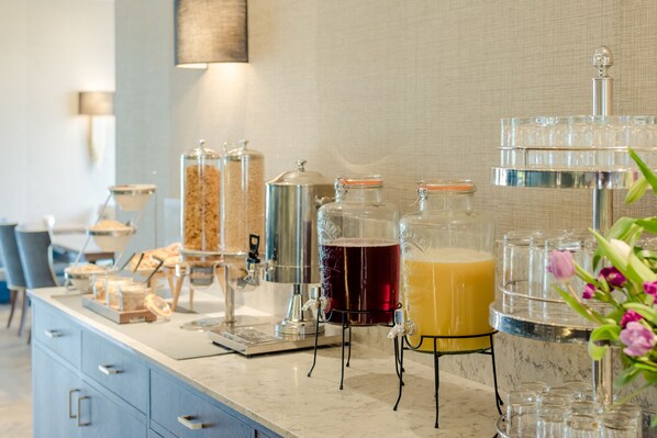 Una barra de desayuno con diversas opciones de comida y bebidas, incluyendo una máquina de café, recipientes de cereal y un dispensador de zumo de frutas.