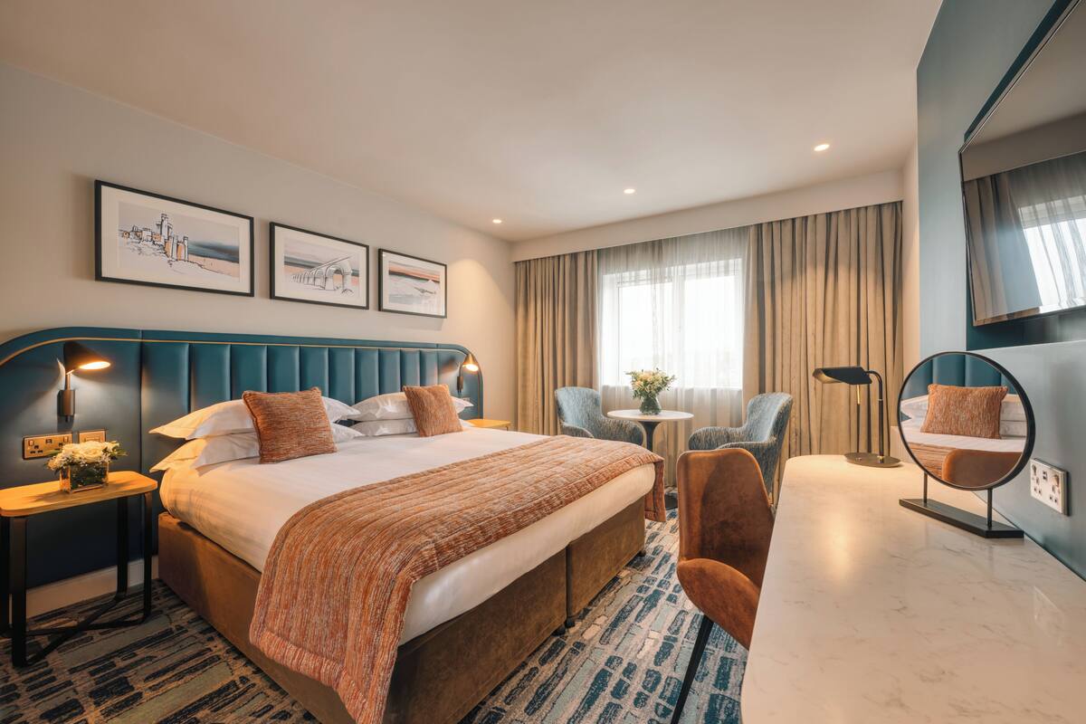 Une chambre d’hôtel avec un grand lit, un miroir rond, un coin salon avec des fauteuils et des œuvres d’art accrochées au mur.