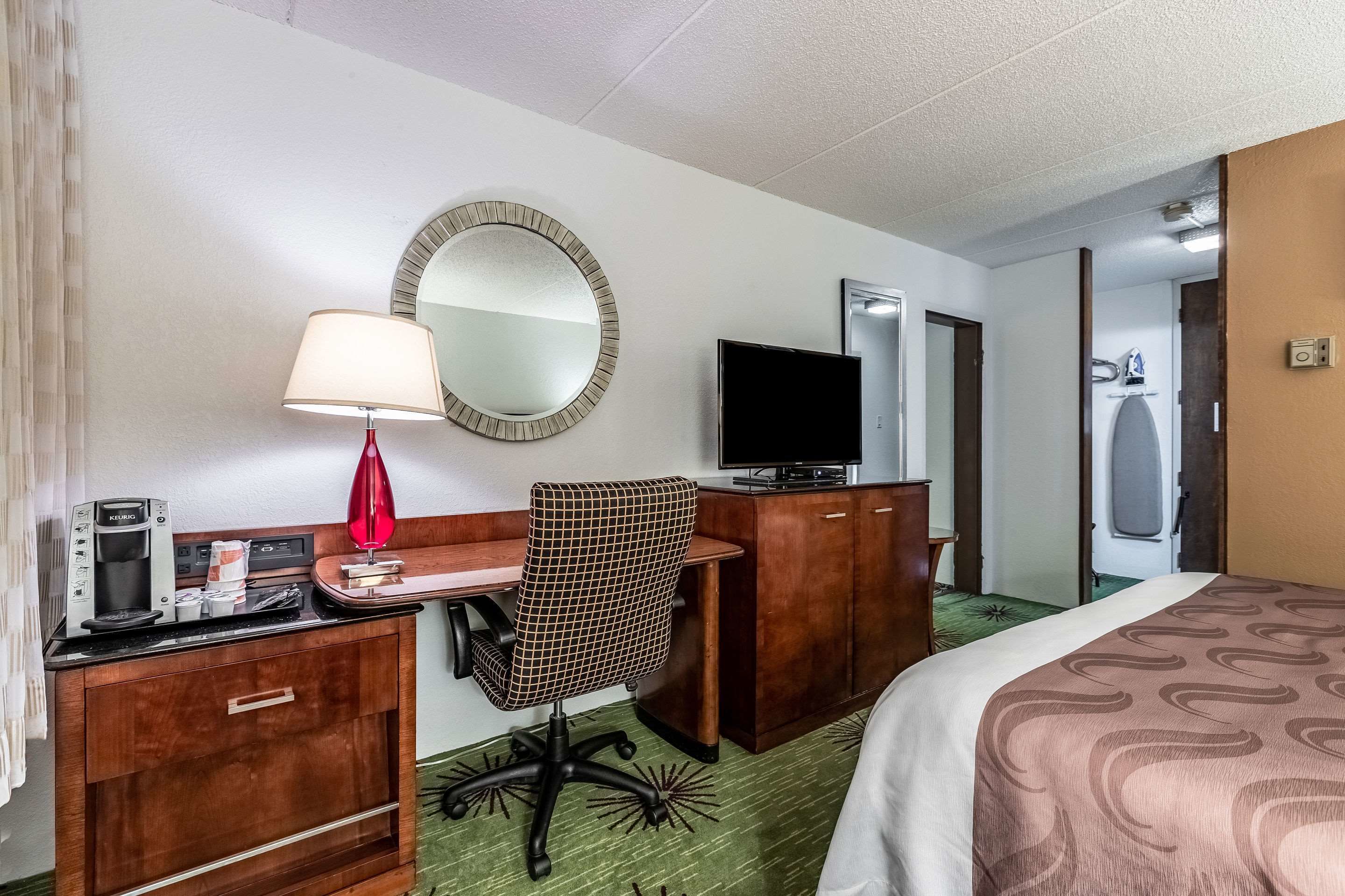 A hotel room with a bed, a desk with a lamp, a television, a mirror, and a wardrobe.