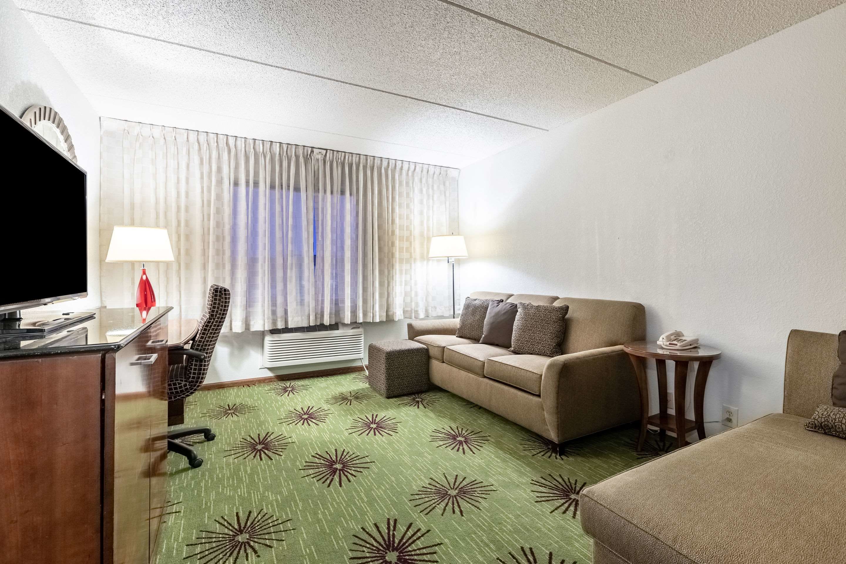 Una habitación de hotel con sofá, una silla, una mesita, televisor y ventana con cortinas.
