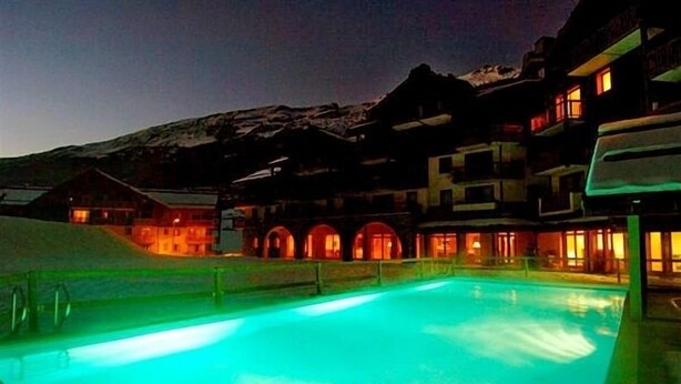 Piscina com montanhas ao fundo, rodeada por edifícios com janelas iluminadas.