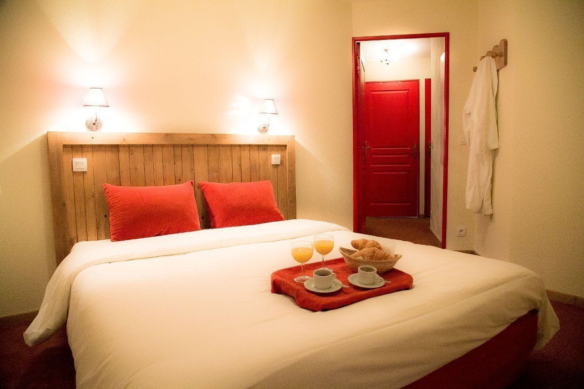 Una cama bien tendida con almohadas rojas, un cesto con alimentos para el desayuno y una puerta roja al fondo.