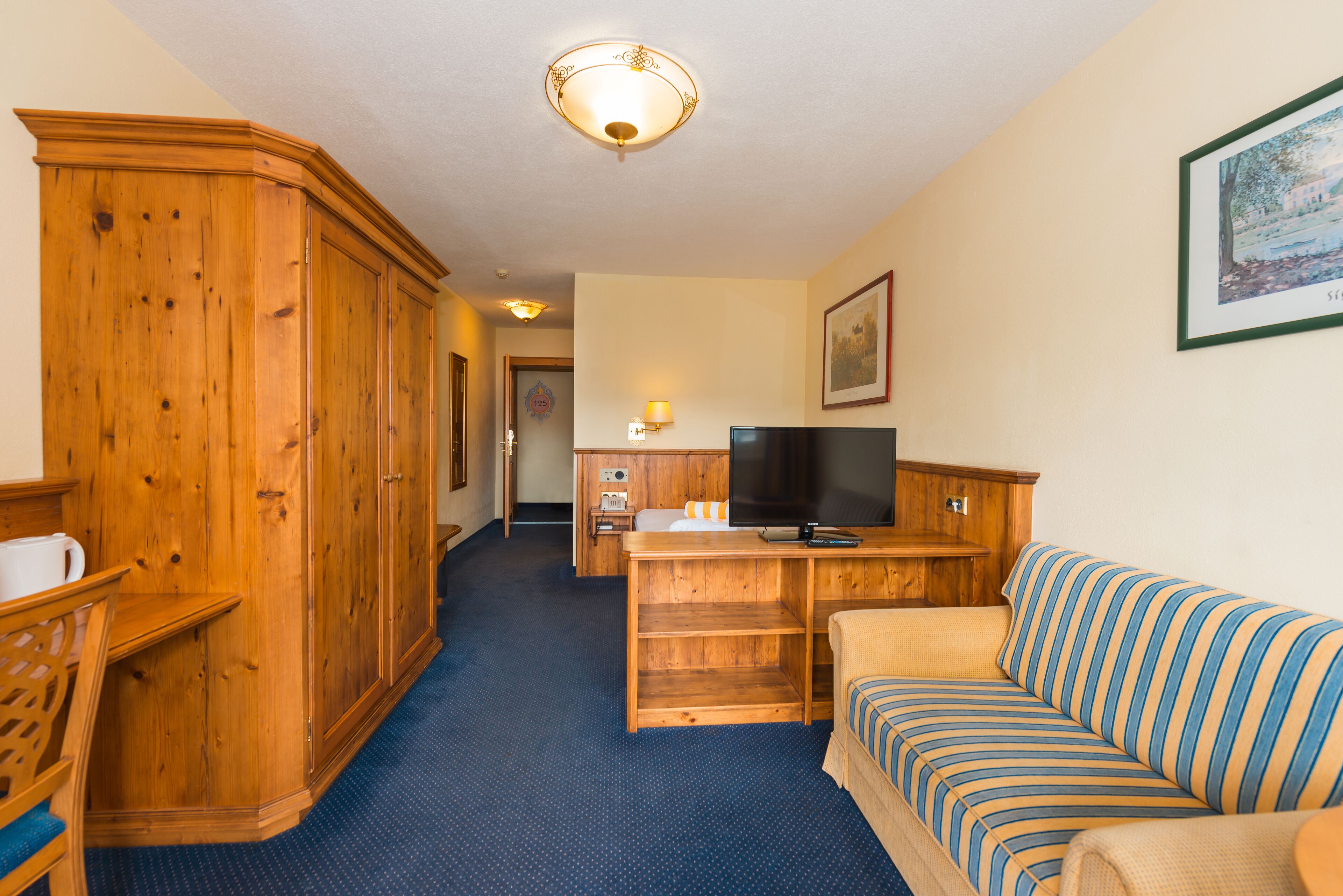 A hotel room with a wooden wardrobe, a striped sofa, a television, and a desk with a lamp.