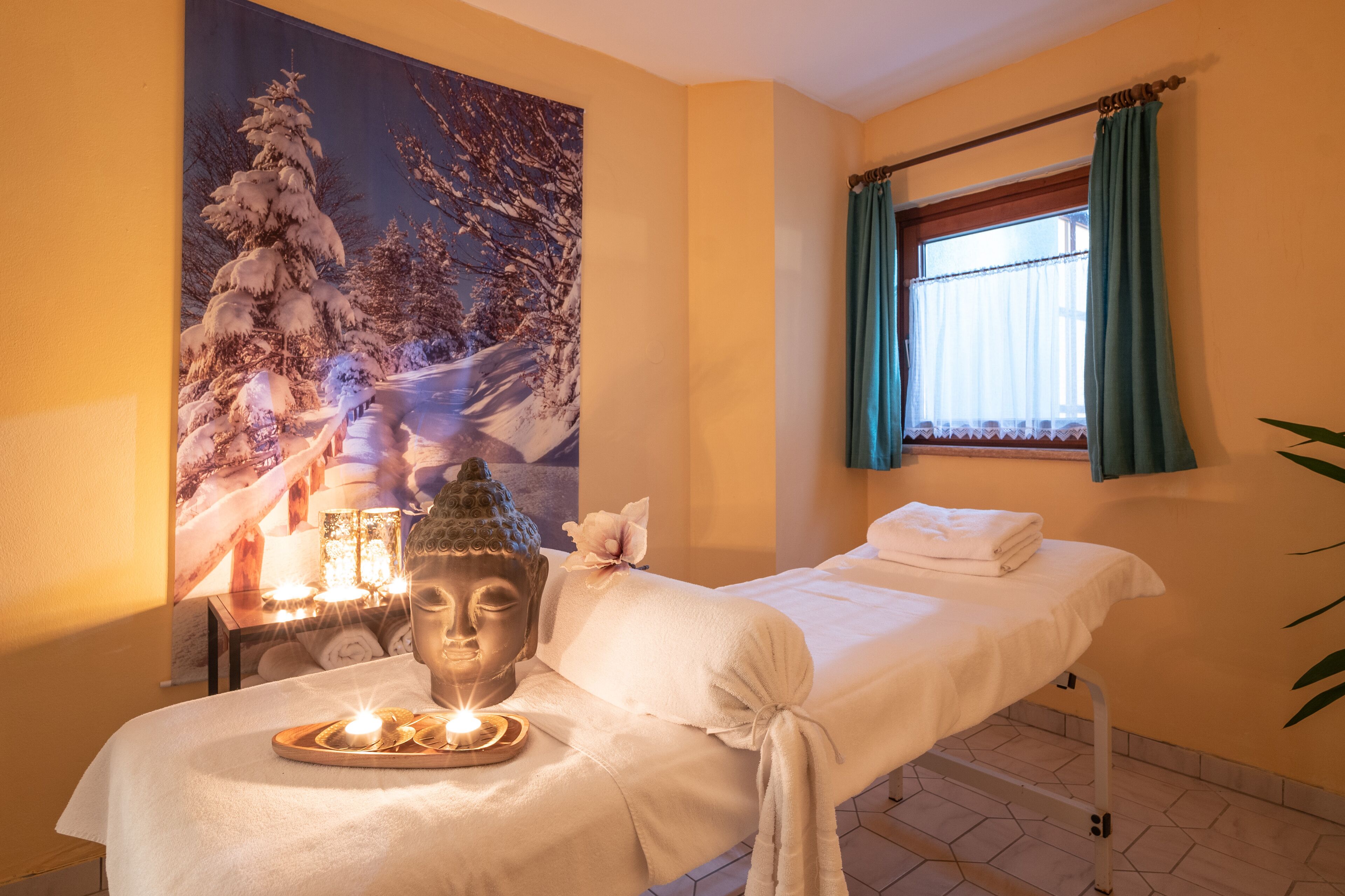 Un cuarto de spa con una camilla de masaje, velas, una estatua de Buda y una pintura con temática invernal.