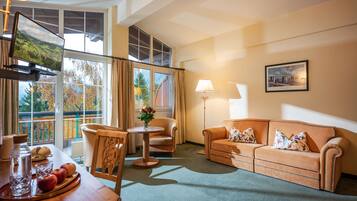 A hotel room with a sofa, armchairs, a dining table with apples and a vase of flowers, a TV, and a window with a view of trees.