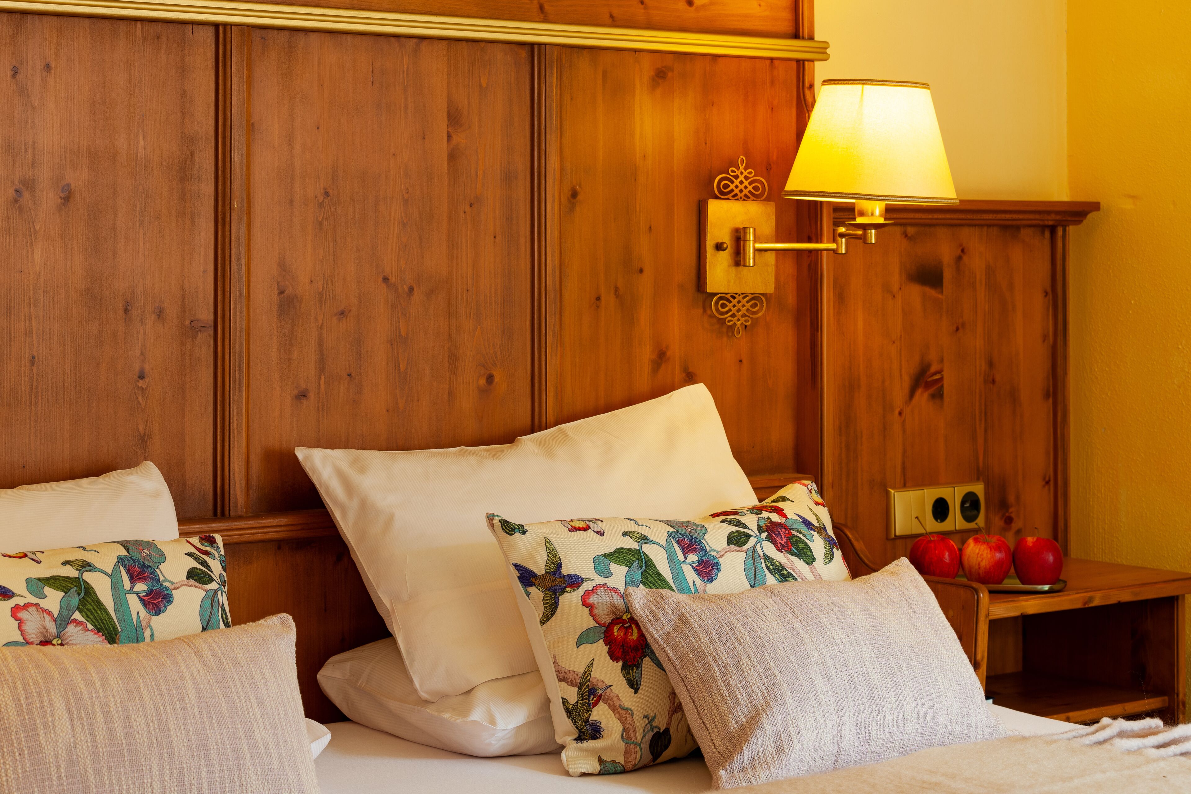 Ein Hotelzimmer mit einem hölzernen Kopfteil, einem Bett mit weißen Leinen, blumigen Kissen und einem Nachttisch mit Äpfeln, sowie einer Wandlampe.