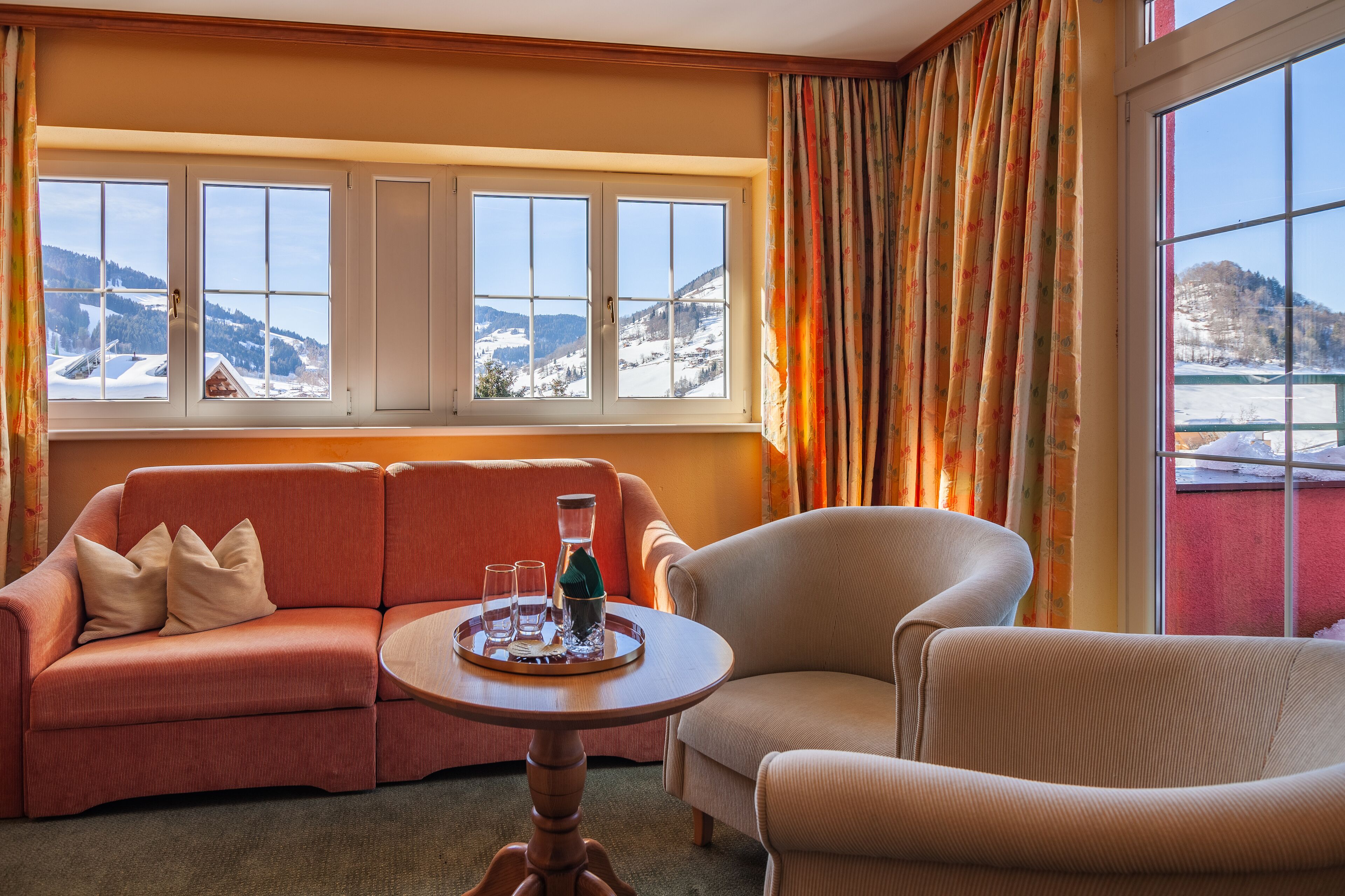 A cozy room with a sofa and armchair, a small table with glasses and a bottle, and a window with a snowy mountain view.