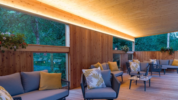 A modern outdoor seating area with wooden flooring and built-in benches, surrounded by large windows offering a view of the greenery outside. - Hotel Sonnschein (Hopfgarten im Brixental)