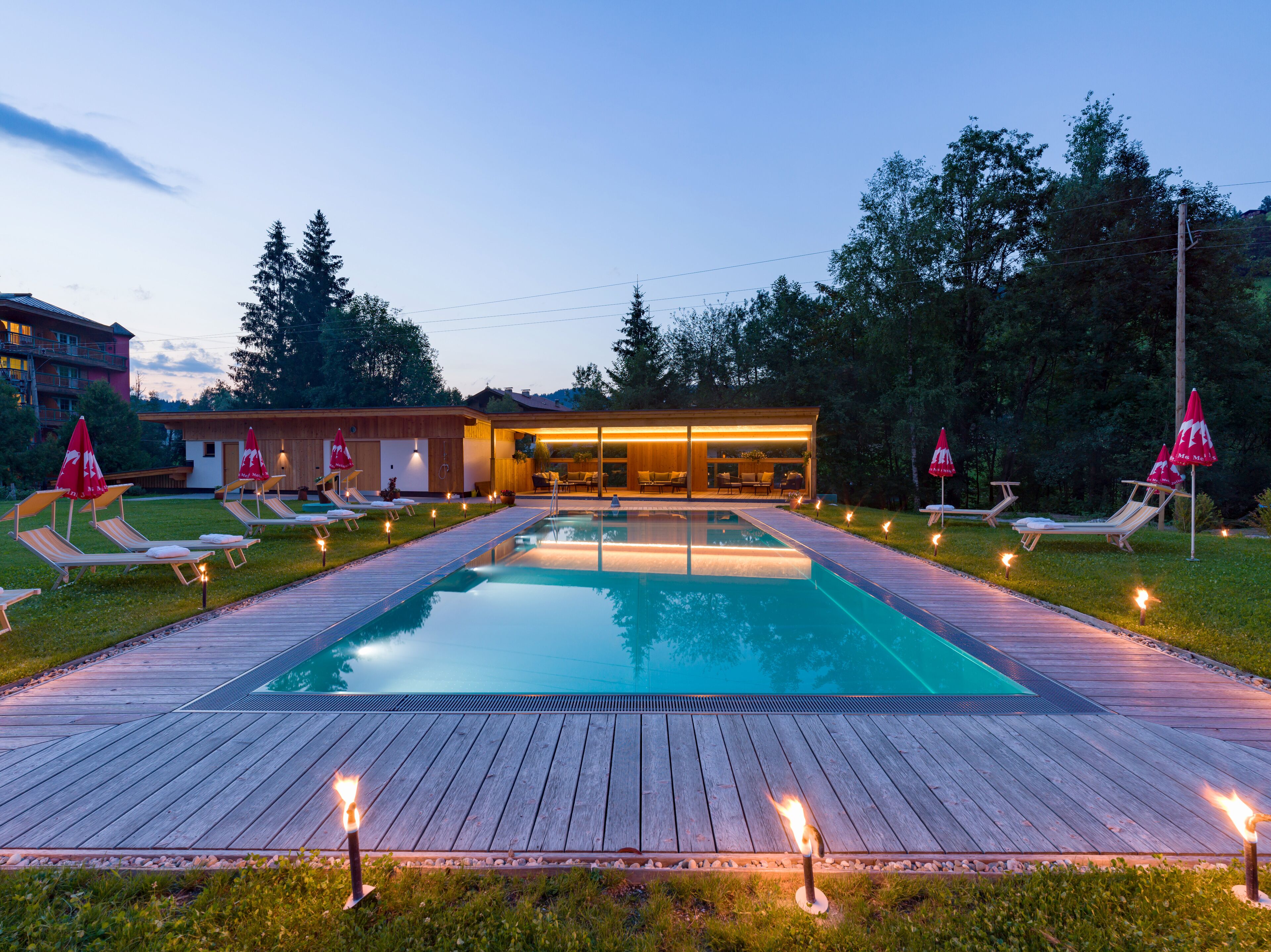 Une piscine entourée de chaises longues et de parasols, elle-même bordée d’une terrasse en bois et d’un bâtiment doté de grandes fenêtres.