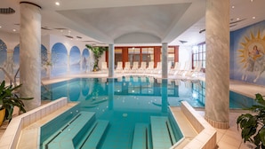An indoor swimming pool with clear water, surrounded by white columns and a sun mural on the wall. - Hotel Sonnschein (Hopfgarten im Brixental)