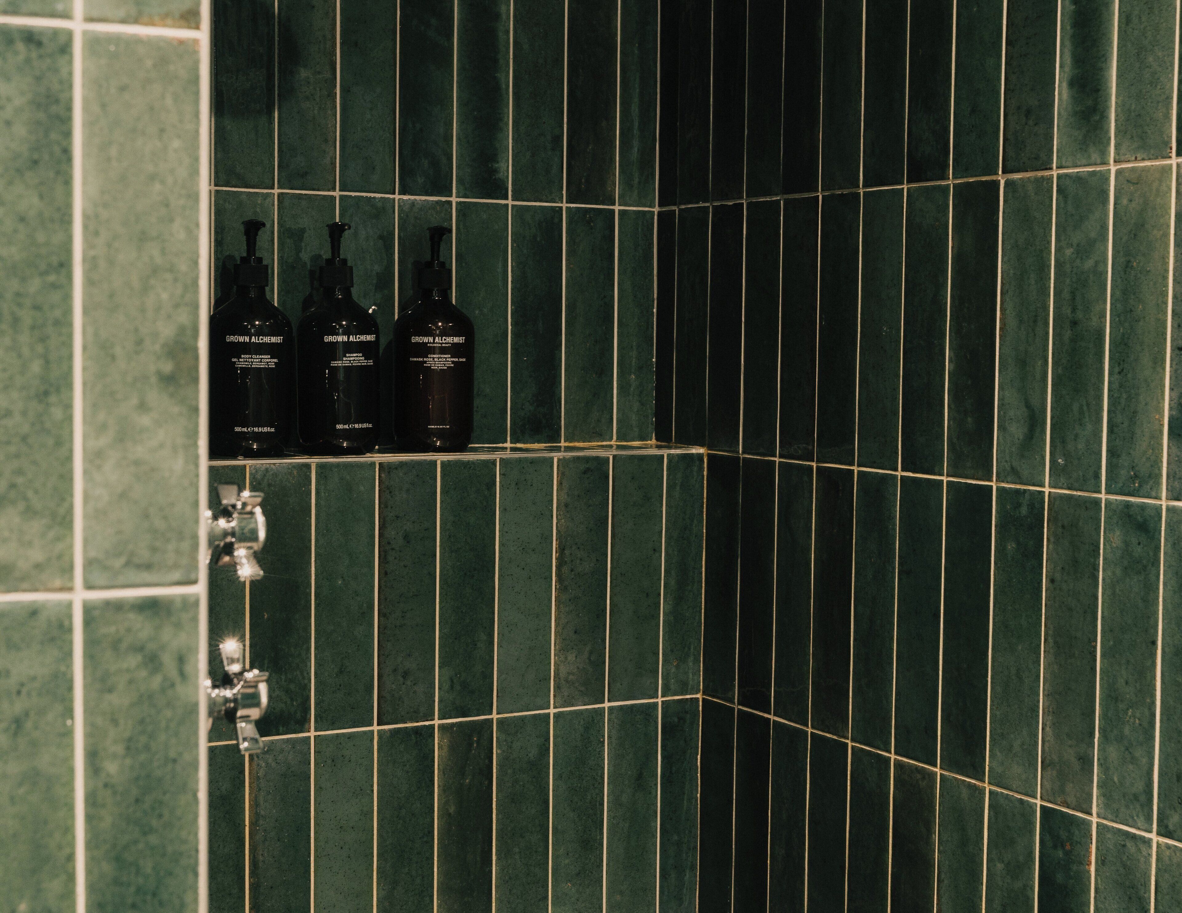 A bathroom with green tiles and three bottles of 'Grown Alchemist' shampoo.