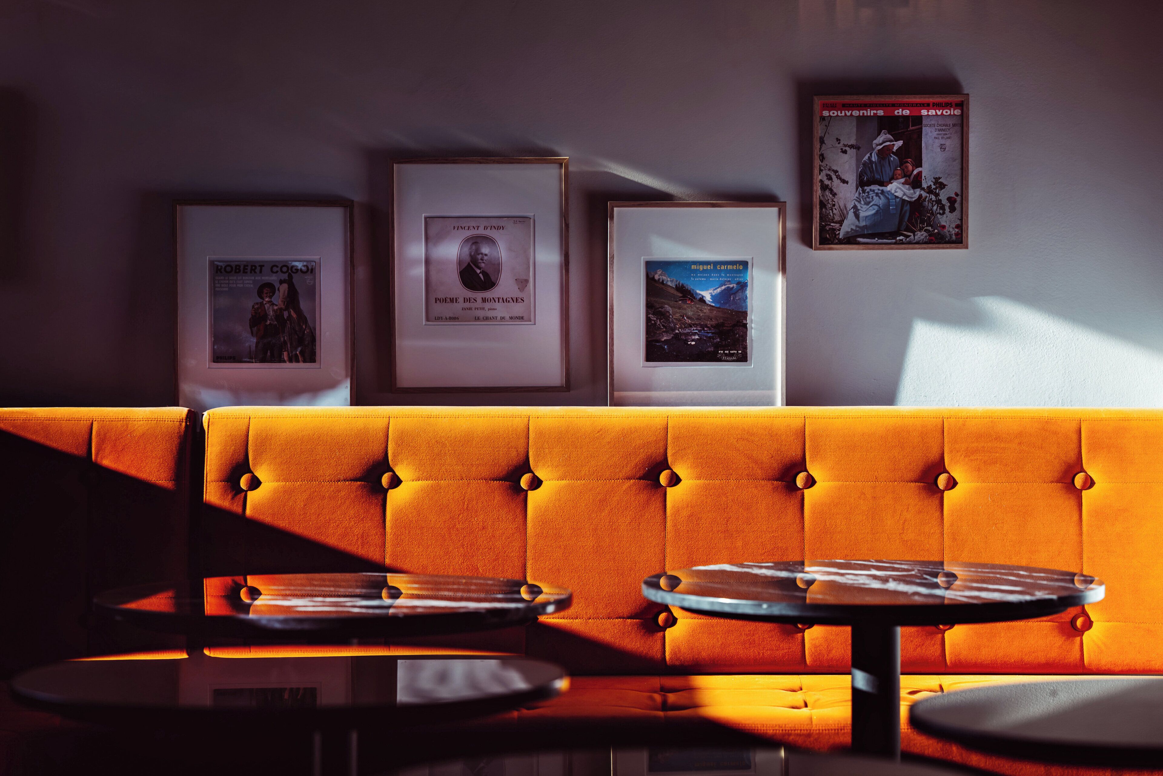 A seating area with a tufted orange sofa and a table, framed artwork on the wall, and a window casting light.
