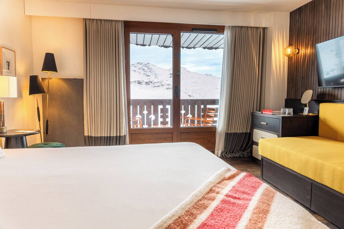 A hotel room with a bed, a desk, a TV, and a view of mountains through the door.