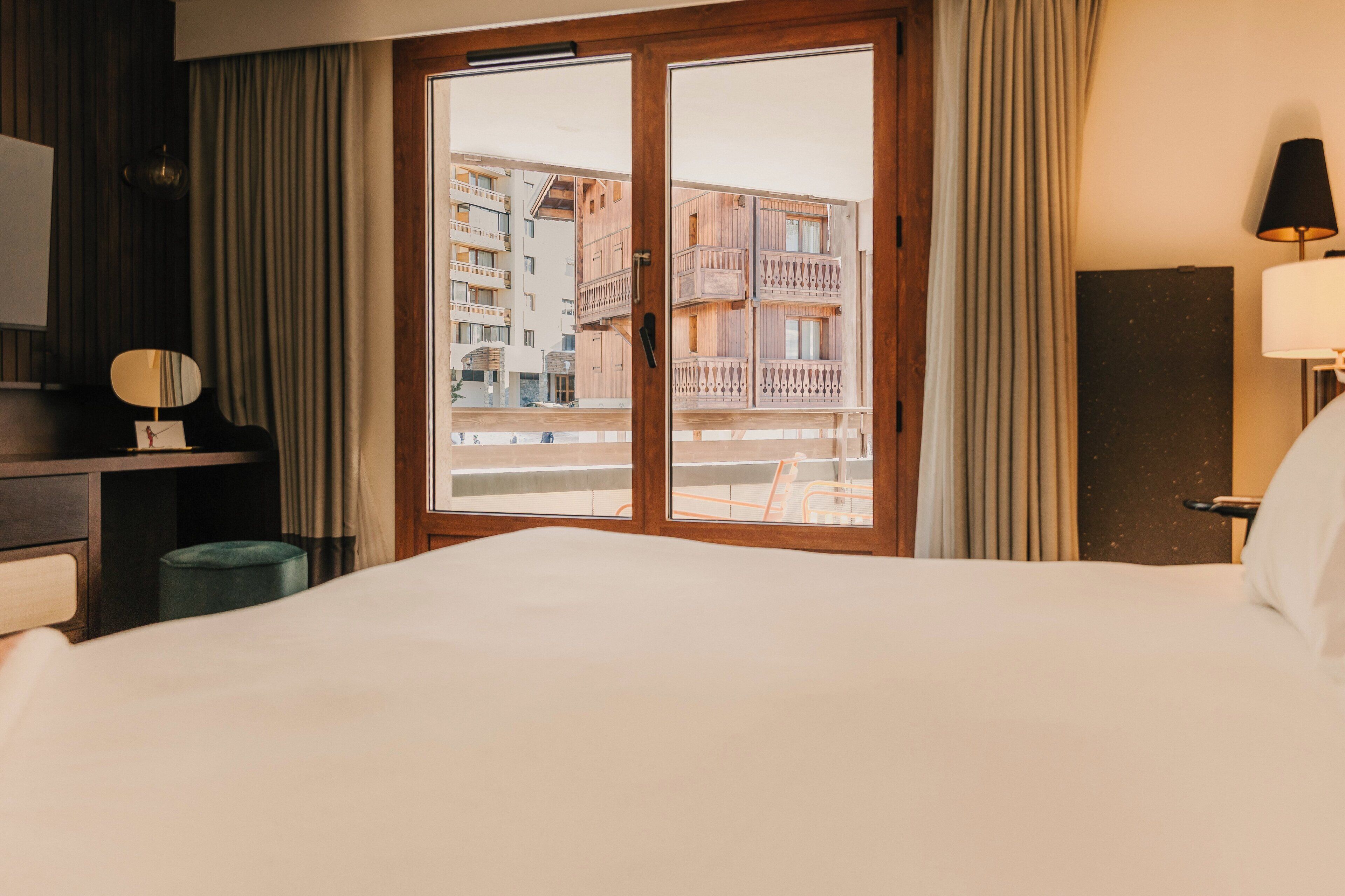 Hotellihuone, jossa on suuri sänky, työpöytä tuolilla, pieni pyöreä pöytä, peili ja ikkuna, josta avautuu näkymä parvekkeelliseen rakennukseen.