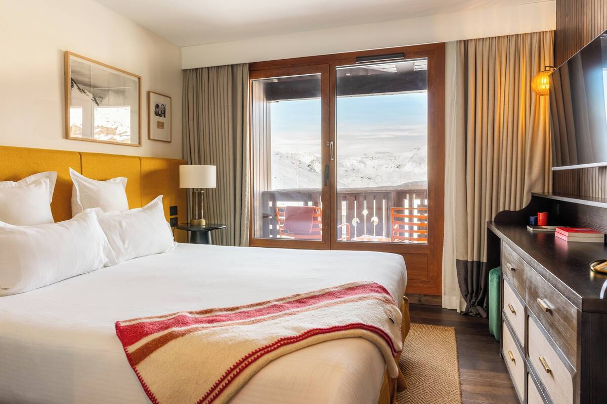 A hotel room with a large bed, a wooden headboard, a desk with a lamp, a view of a snowy landscape through a sliding door, and a framed picture on the wall.