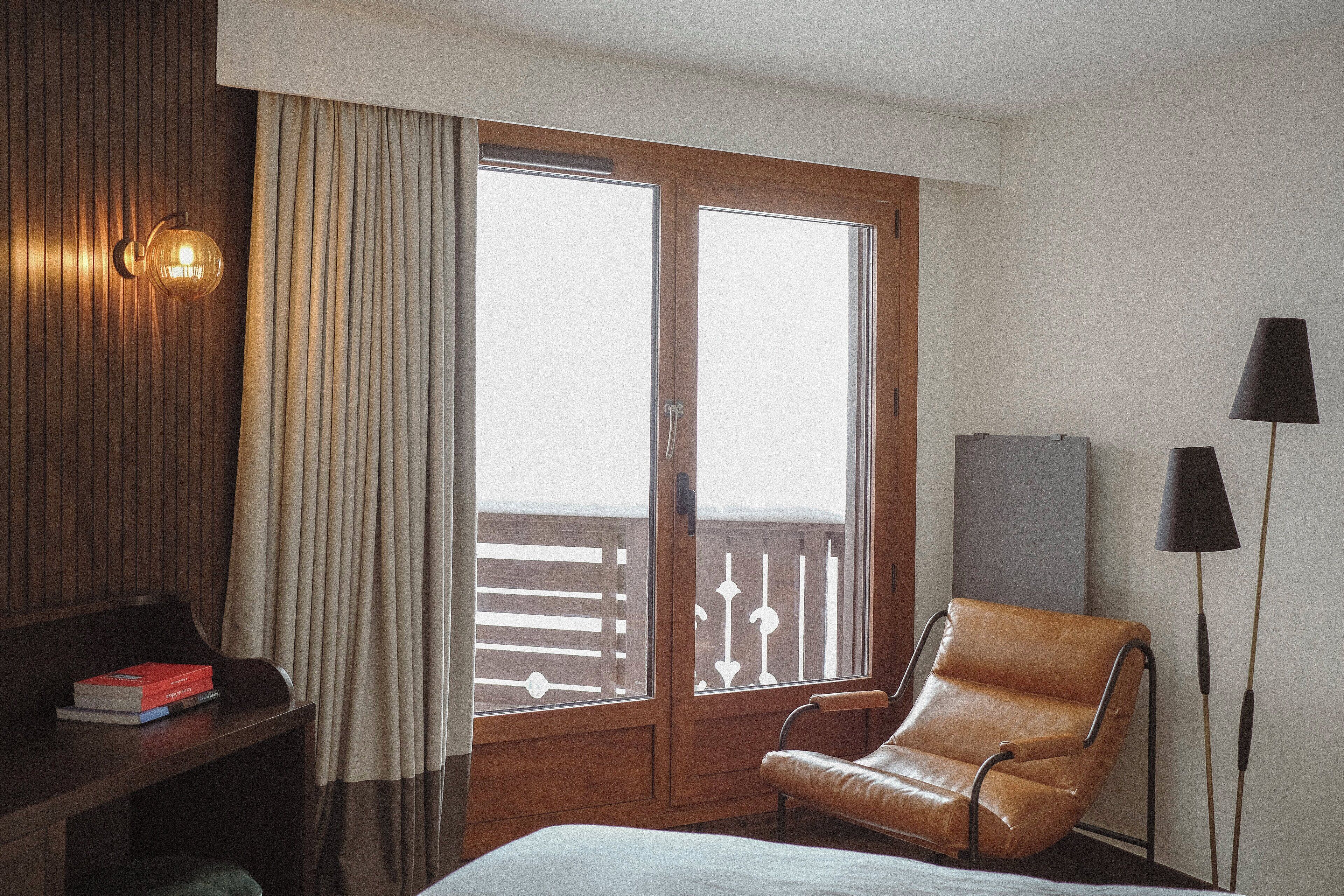 Een hotelkamer met een houten bureau, een lederen fauteuil, een bed, een raam met gordijnen en een lamp.