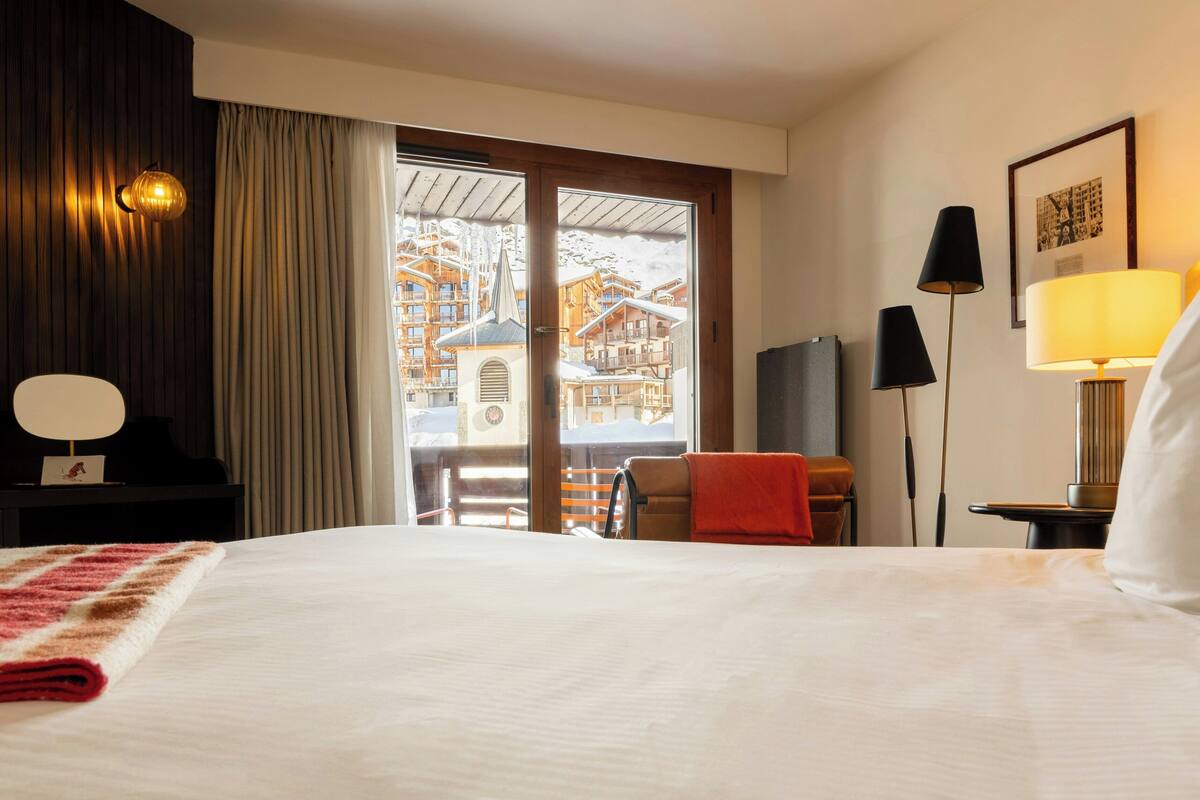 A hotel room with a large bed, a view of a ski resort, a desk with a lamp, and a chair.