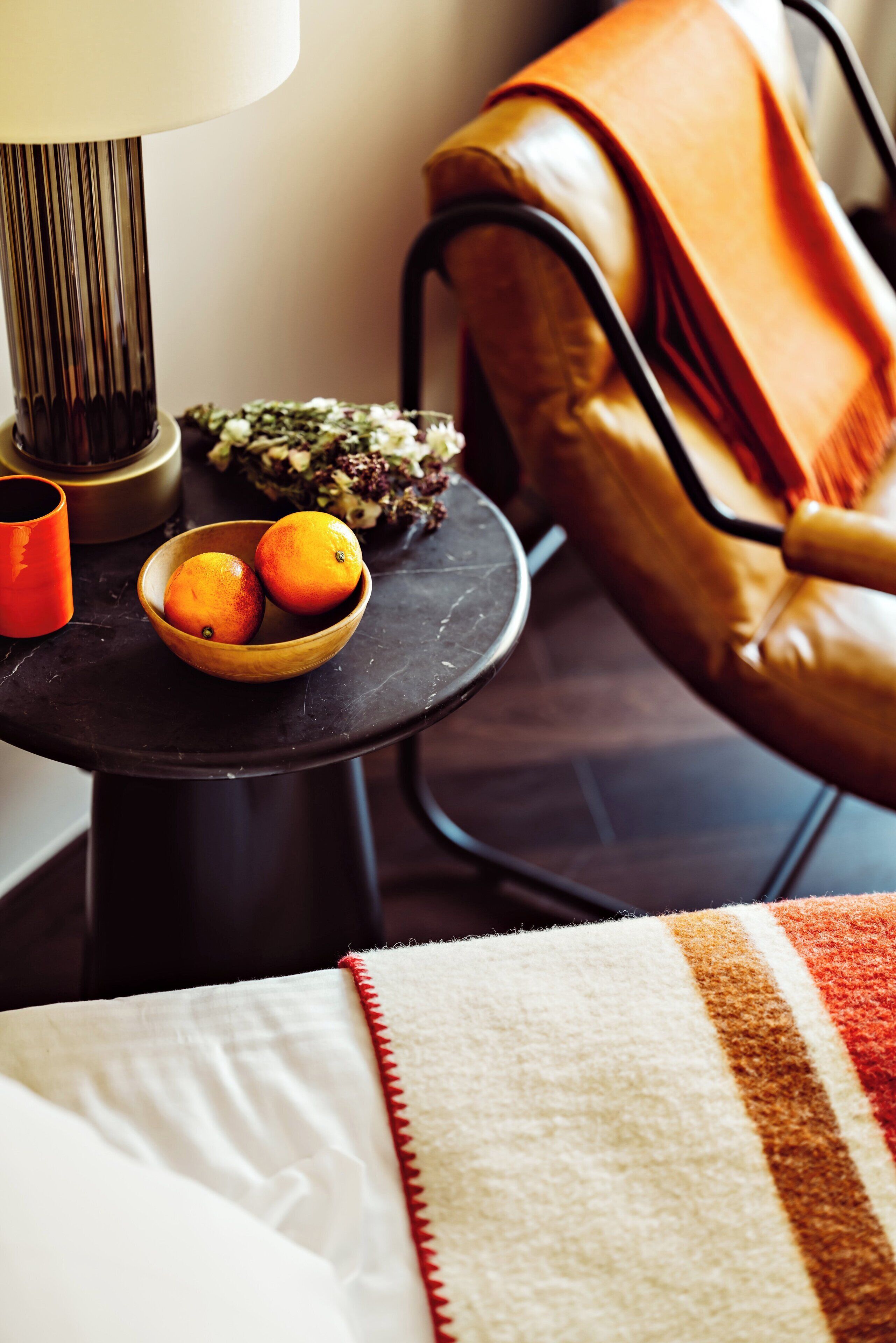 A small round table with a bowl of oranges, a lamp, and a vase with flowers.