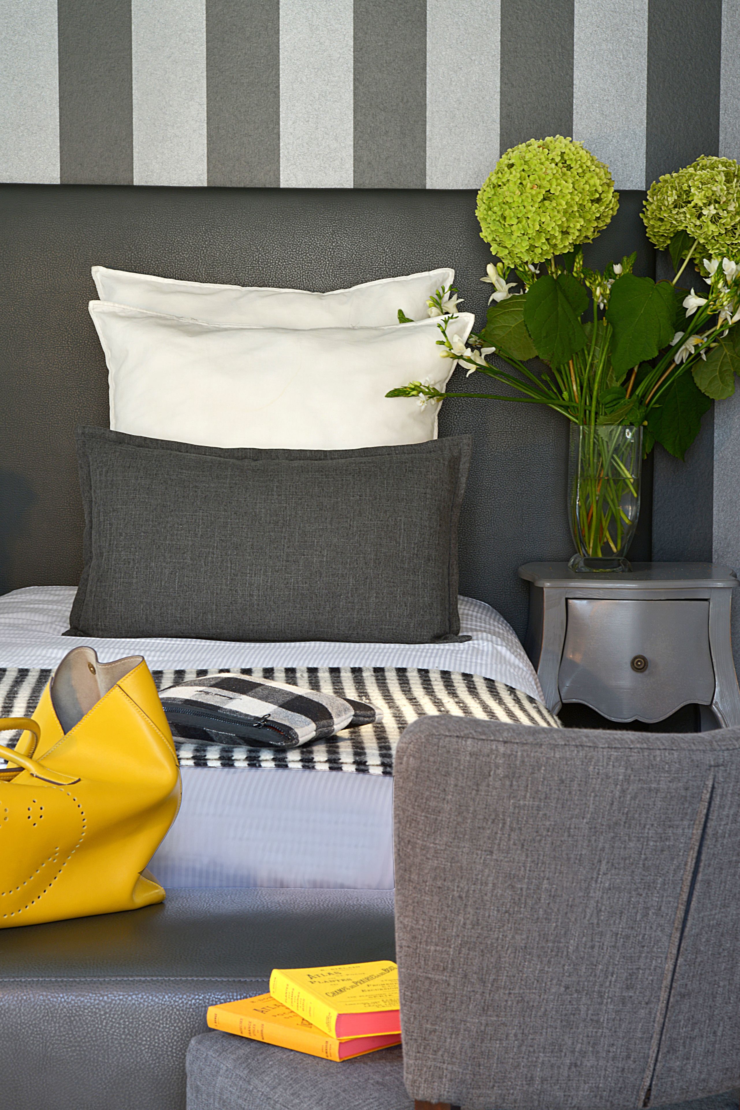 Un dormitorio ordenado con cama, almohadas, una mesita de noche, un florero con flores, un bolso amarillo y un libro.