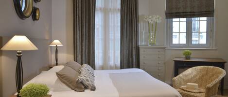 A neatly arranged bedroom with a bed, two bedside lamps, a dresser, a chair, a window with curtains, and a vase with flowers.