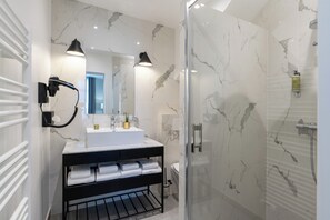 A modern bathroom with a marble shower, a glass-enclosed shower, a white sink with a black countertop, and two wall-mounted lamps. - Hotel Le Fer à Cheval (Trouville-sur-Mer)