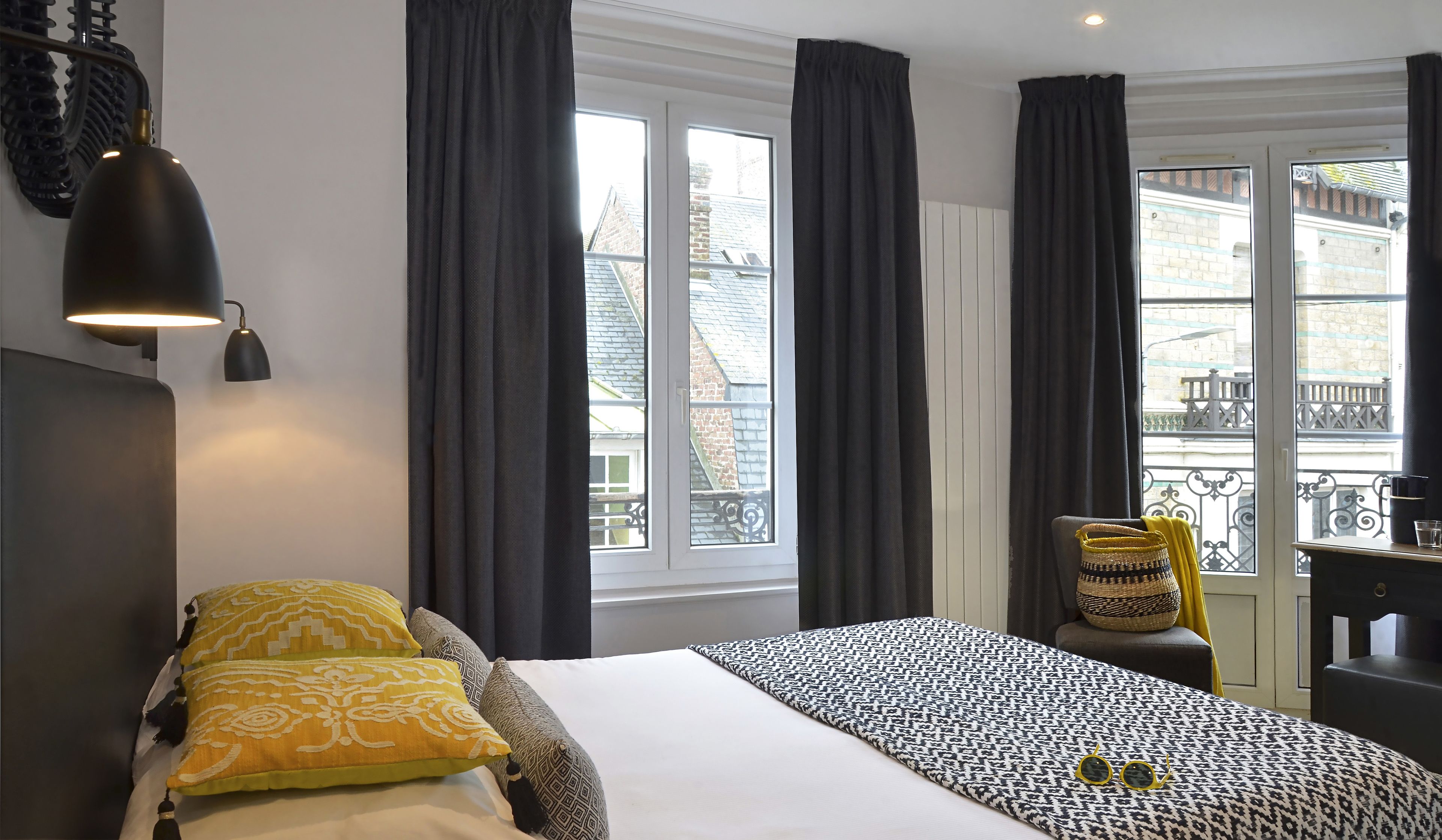 Un dormitorio con cama, cortinas oscuras, una ventana con vista a edificios, una lámpara y un sillón con una cesta.