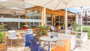 An outdoor cafe area with round tables and chairs, potted plants, and a view of a building with a sign reading "Vista Harbor". - BLUESEA Montevista Hawai (Lloret de Mar)